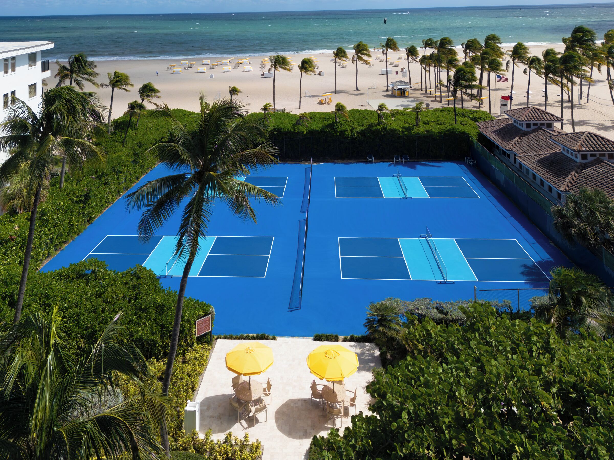 Una cancha de tenis rodeada de palmeras, un área de piscina con sombrillas y una playa al fondo.