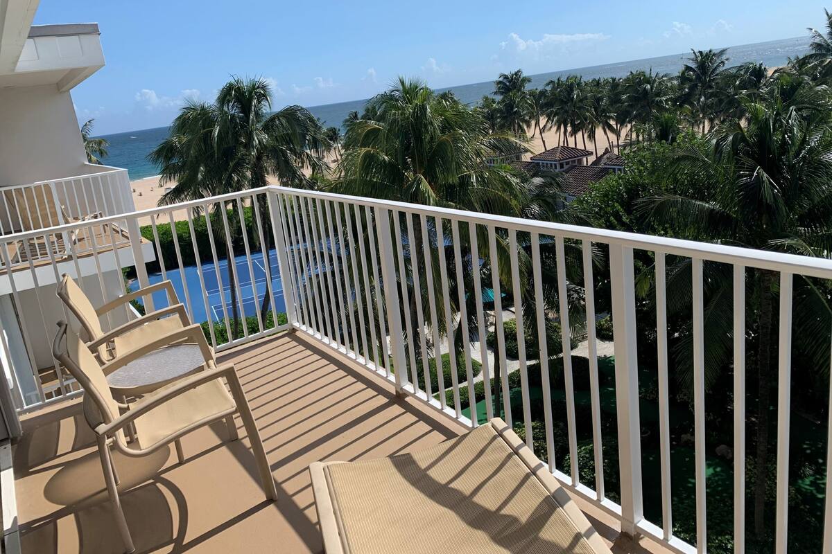 A balcony with a view of a beach, palm trees, and a swimming pool.