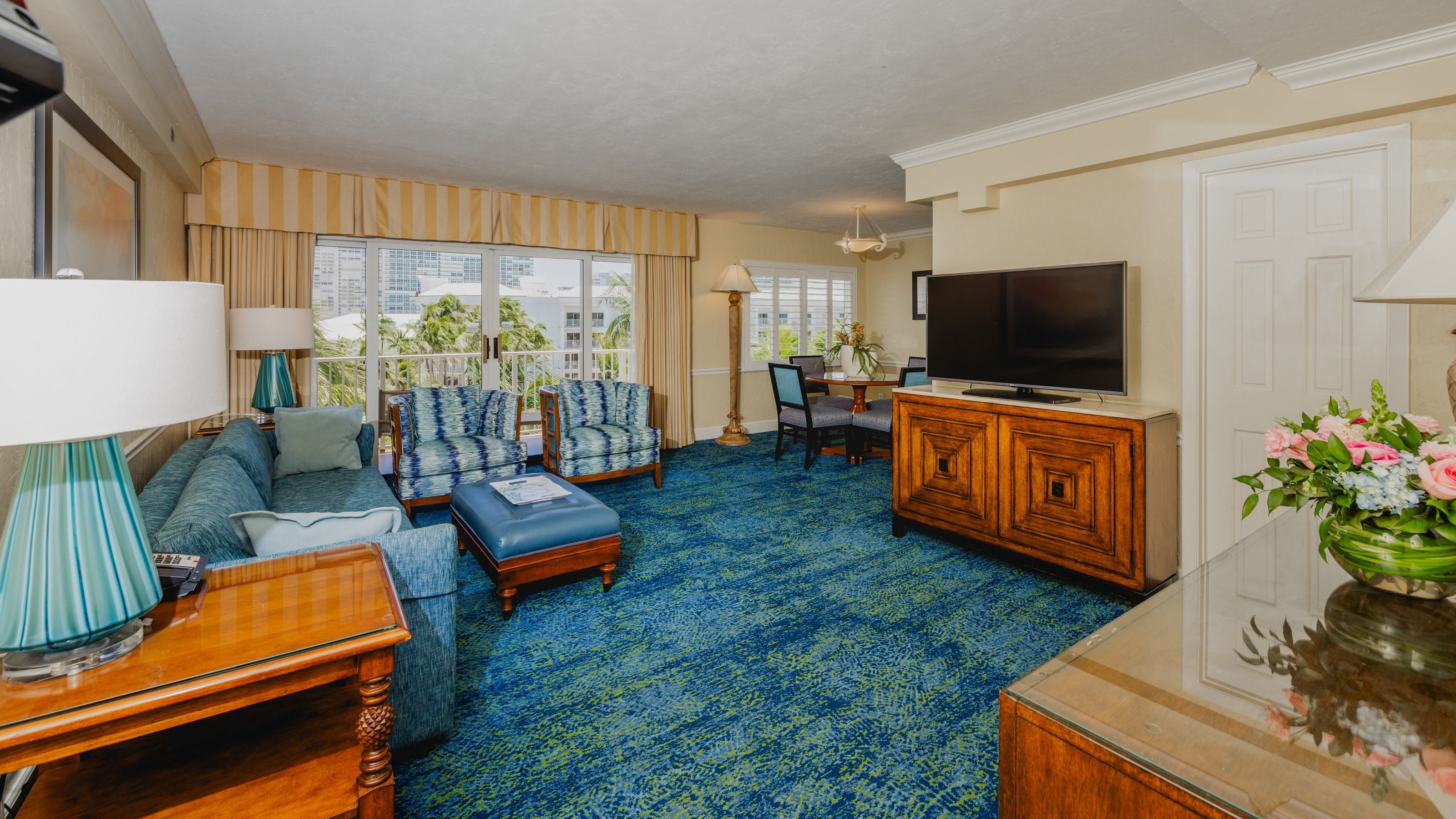 A hotel room with a large TV, a sofa, a dining table, and a balcony with curtains.