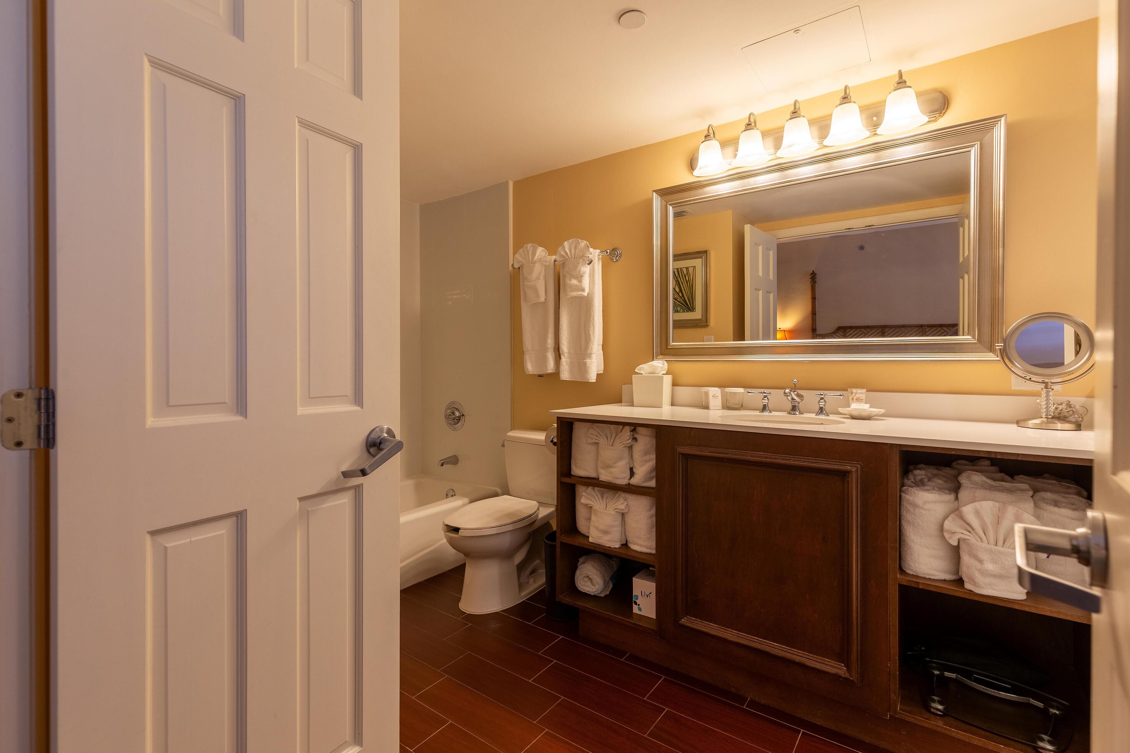 A bathroom with a white toilet, a bathtub, a sink with a mirror, and shelves with towels.