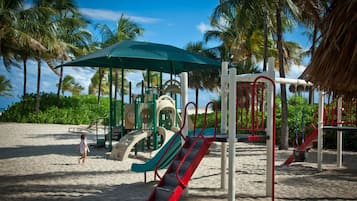 Une aire de jeux de plage avec des toboggans et des structures d’escalade, entourée de palmiers et sous un ciel dégagé.