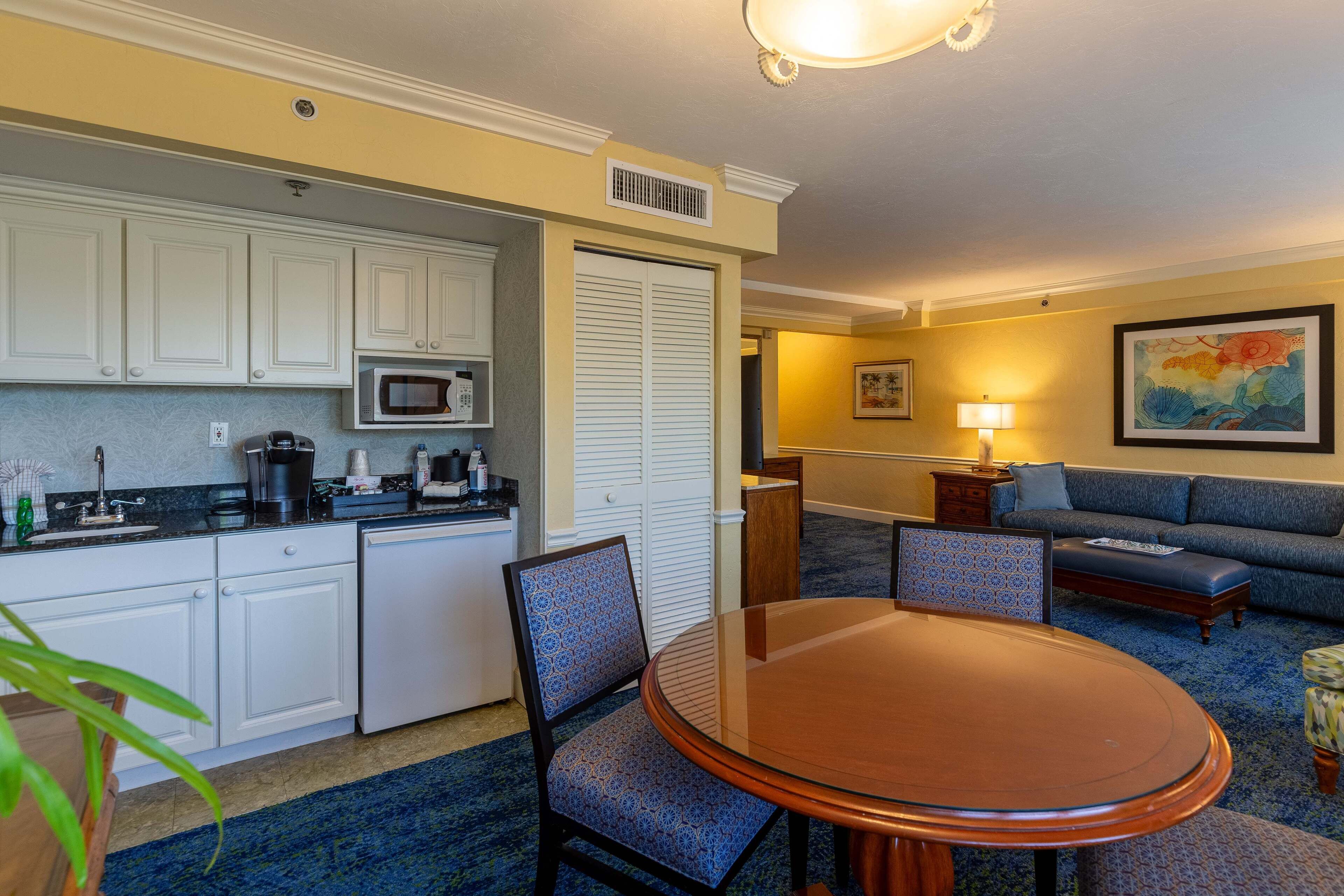 A compact hotel room with a kitchenette, a dining table with chairs, a sofa, and a lamp.