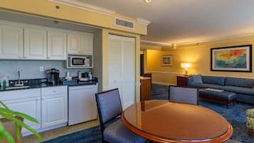 A compact hotel room with a kitchenette, a dining table with chairs, a sofa, and a lamp.