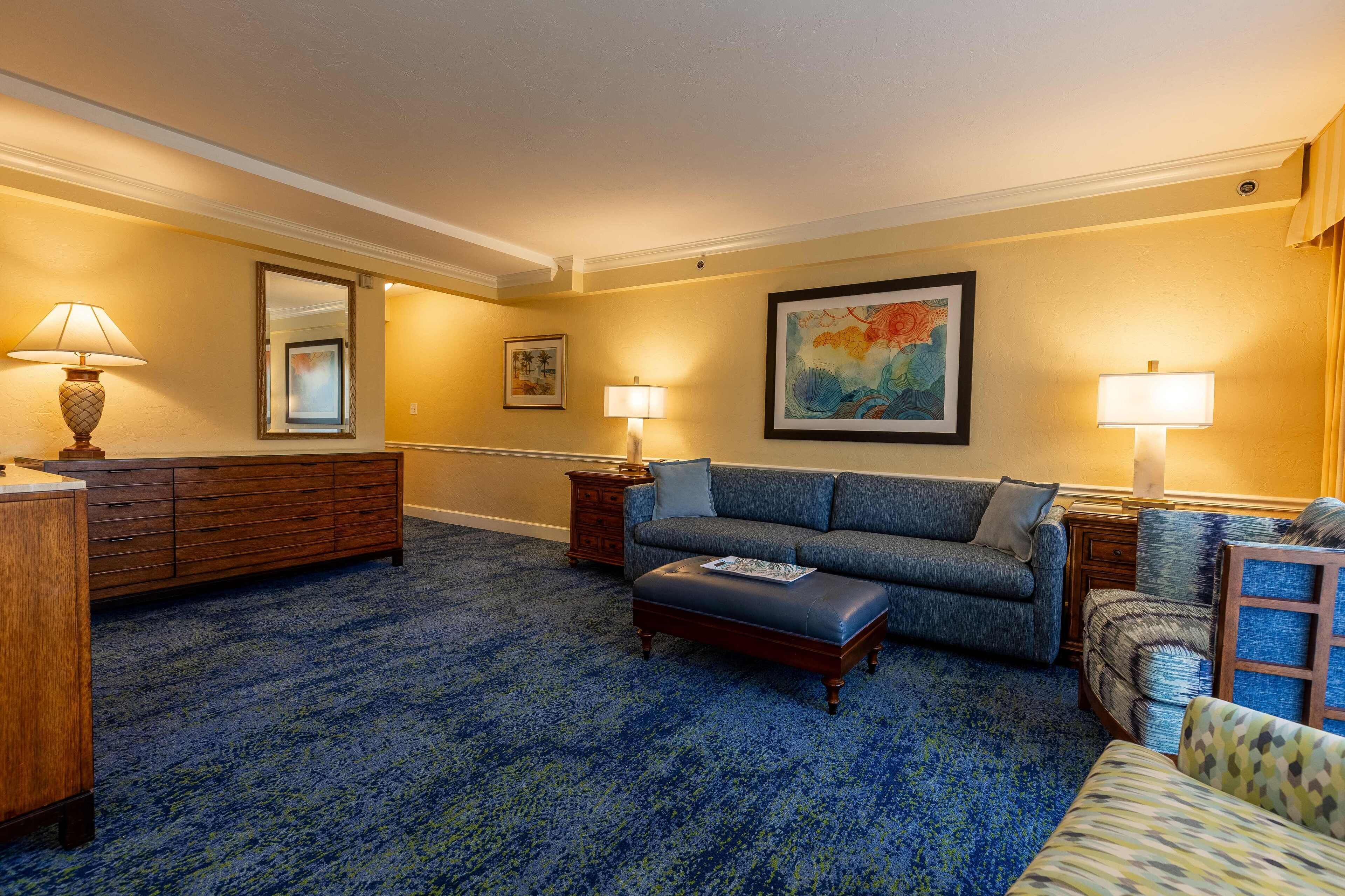 Habitación de hotel con alfombra azul, un sofá, una silla, un aparador de madera y dos lámparas.
