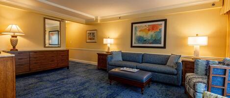 Habitación de hotel con alfombra azul, un sofá, una silla, un aparador de madera y dos lámparas.