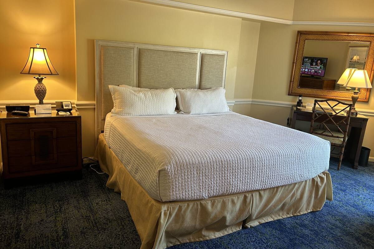 A neatly made bed with a quilted headboard, a bedside table with a lamp, and a mirror on the wall.