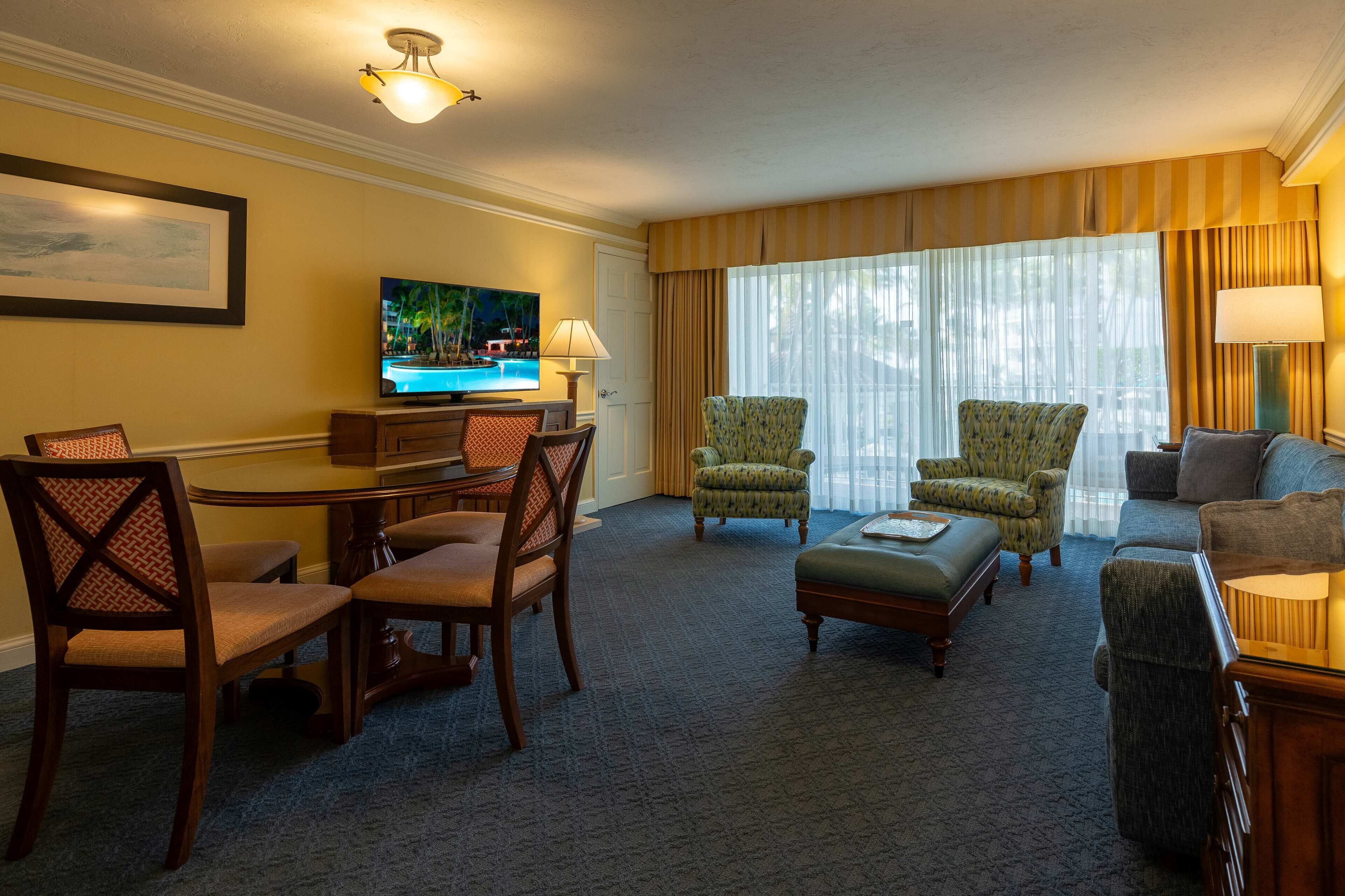 A hotel room with a dining table, two armchairs, a sofa, a TV, and a window with curtains.