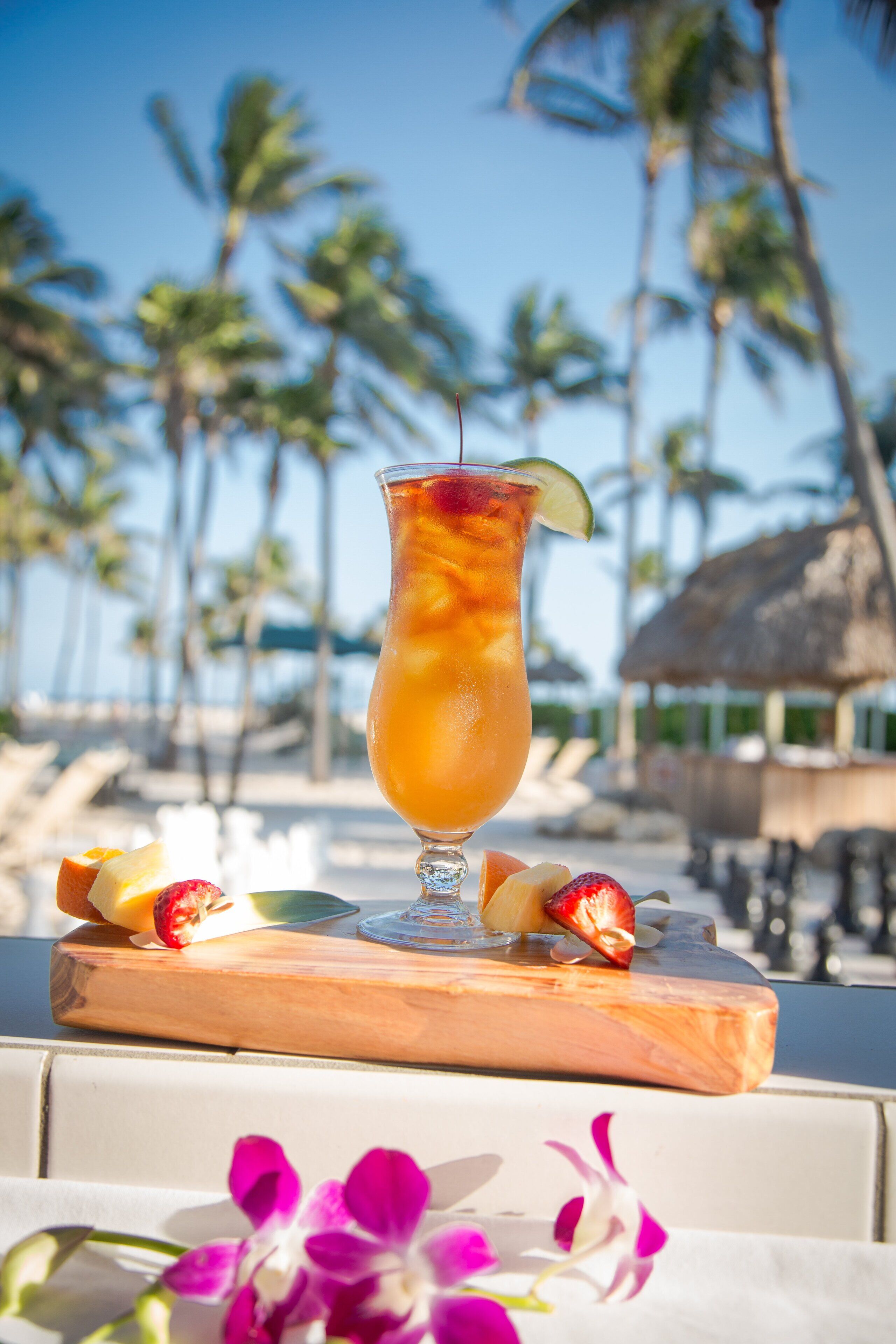 Bebida tropical sobre una tabla de madera con frutas, frente a una playa con palmeras y una estructura de techo de paja.