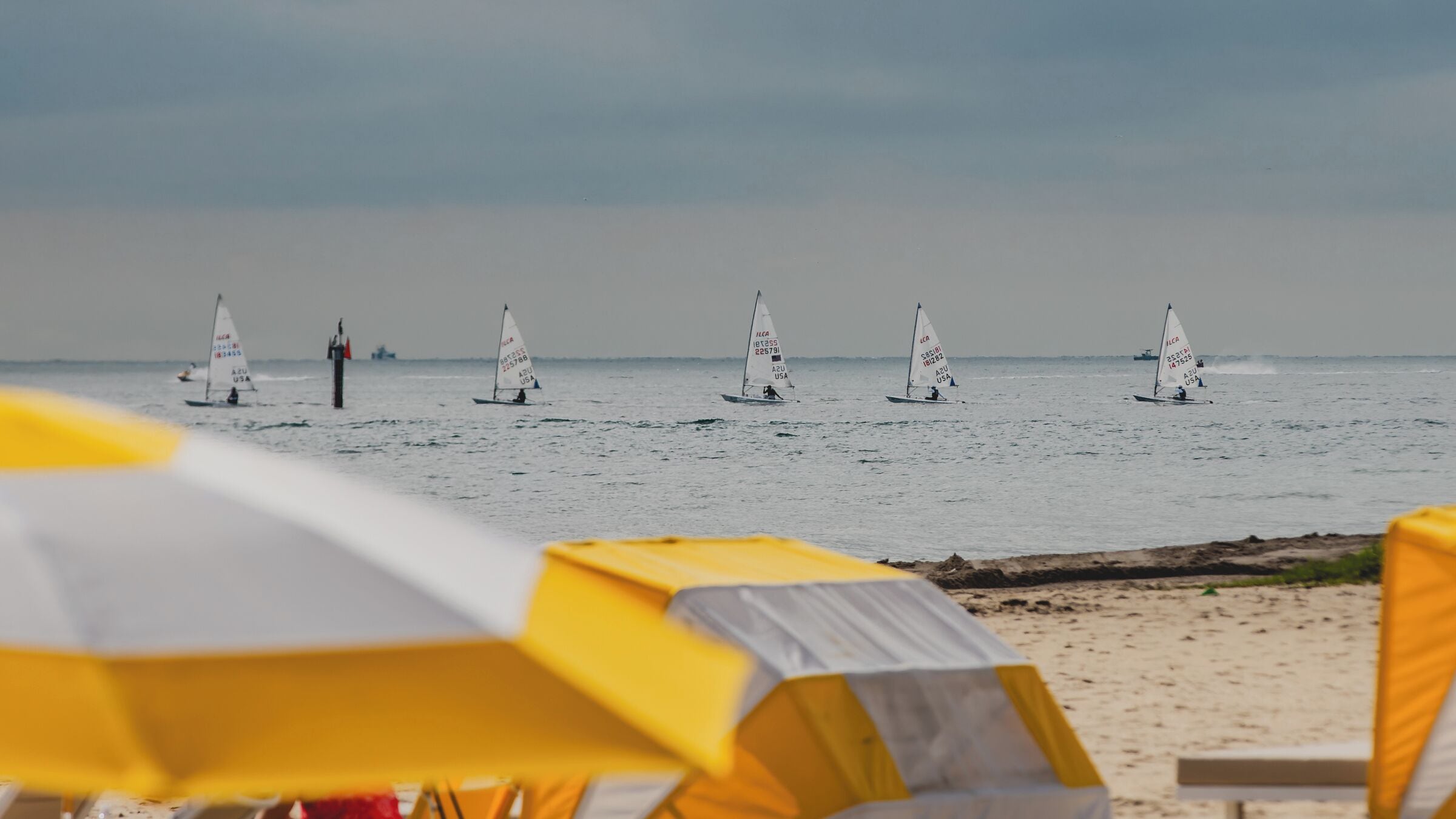 A beach with multiple sailboats and beach umbrellas.