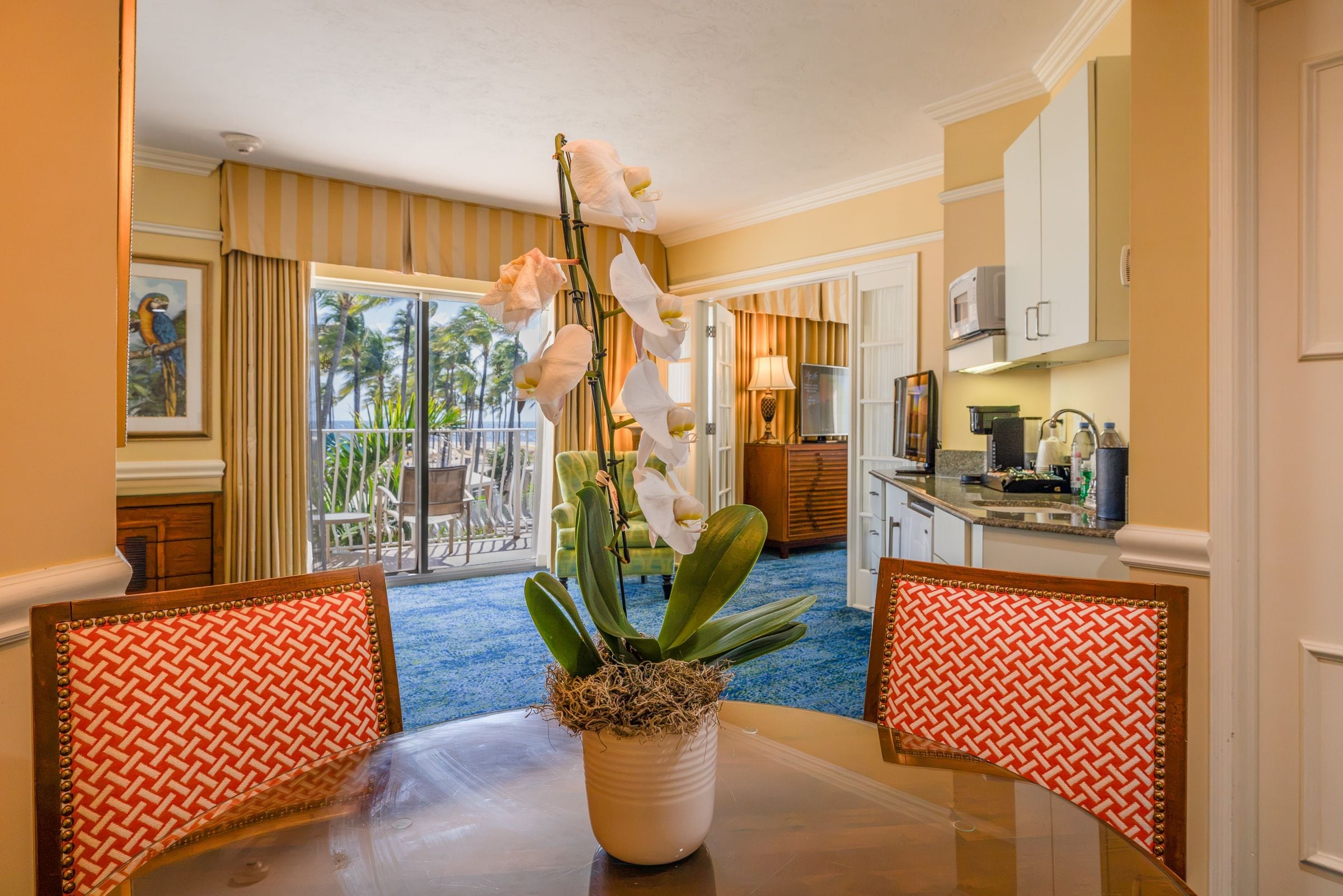 A hotel room with a dining table, chairs, a potted orchid, and a view of the beach through the window.