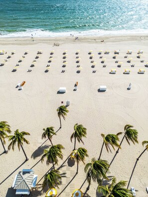 A beach with rows of sun loungers and umbrellas, palm trees, and a lifeguard tower. - Lago Mar Beach Resort & Club (Fort Lauderdale)