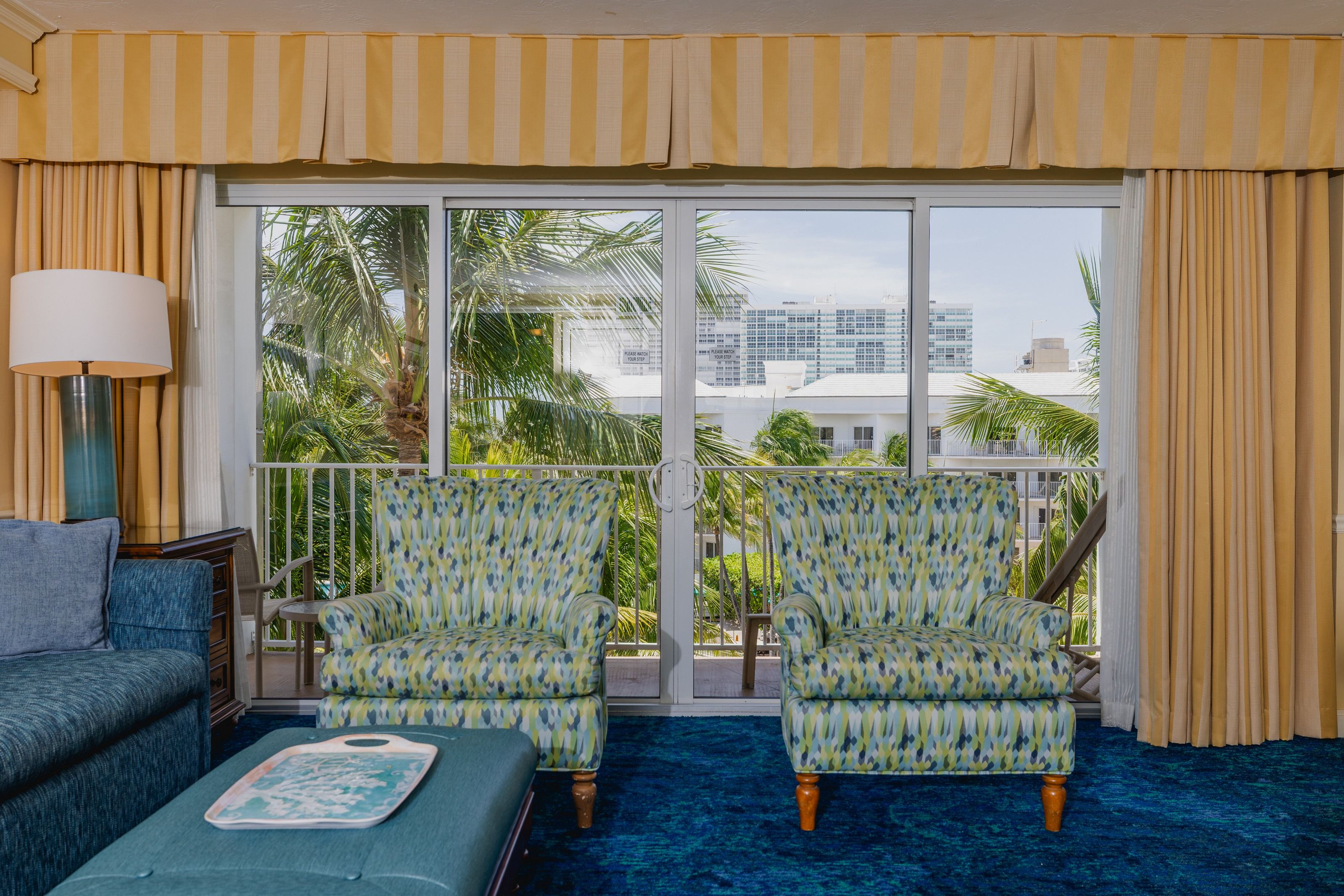 Une chambre d’hôtel avec un balcon, deux fauteuils à motifs, un canapé, une lampe et une vue sur des palmiers et des immeubles.