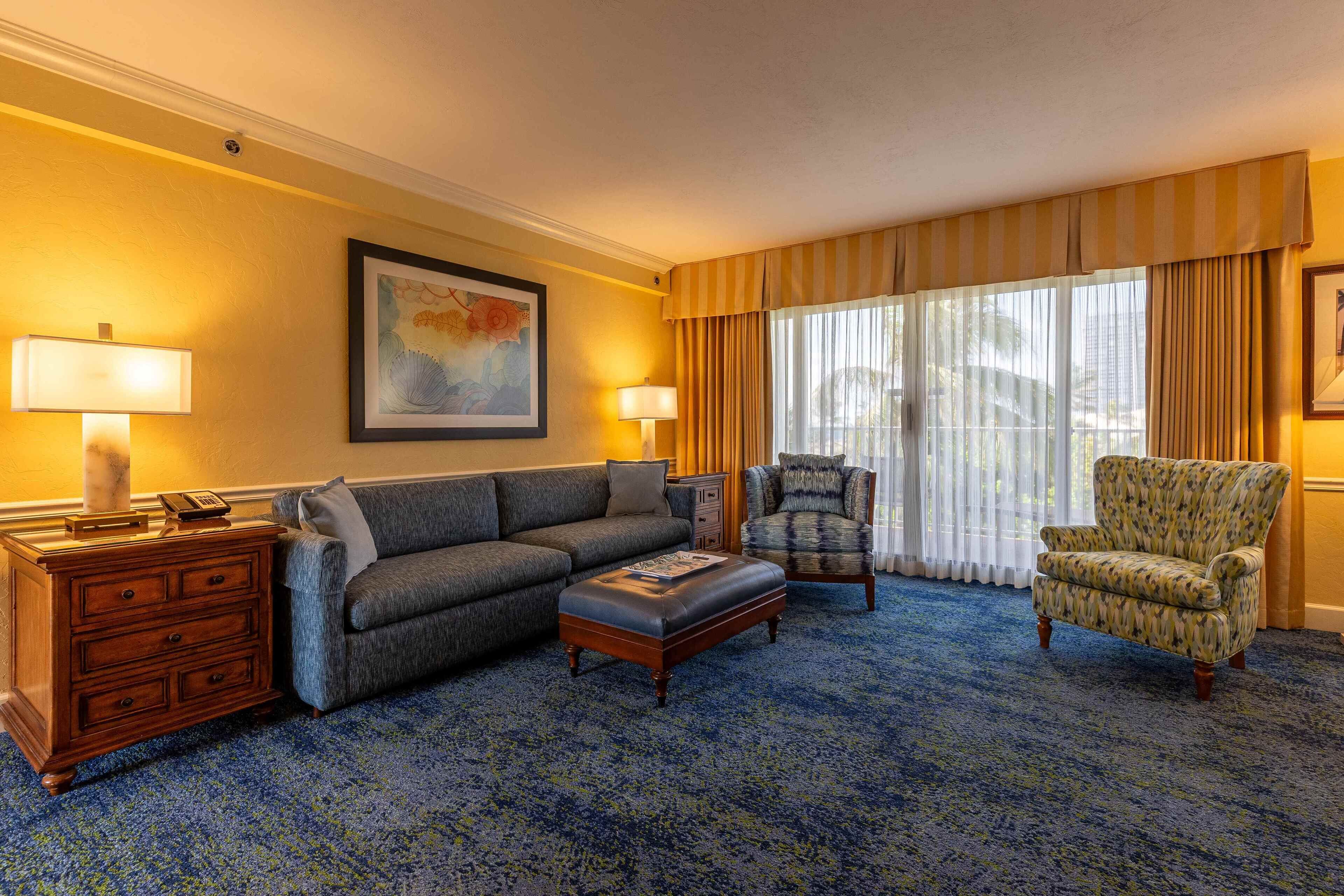A hotel room with a sofa, armchair, ottoman, wooden dresser, and a large window with curtains.