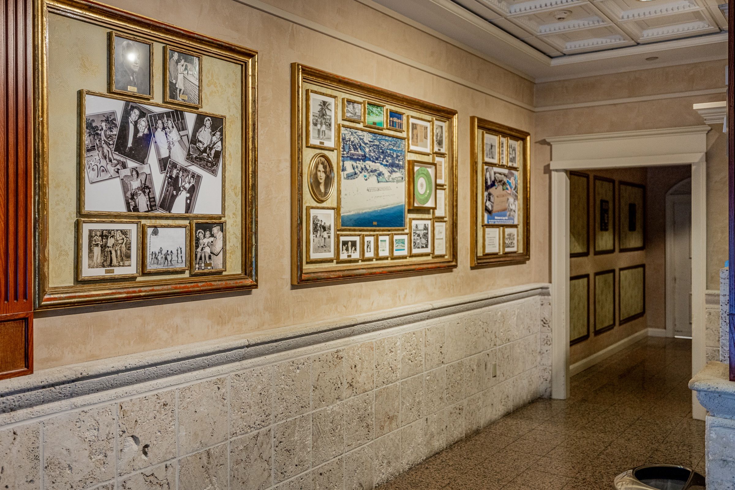 A hallway with framed photographs on the wall, a tiled floor, and a door at the end.