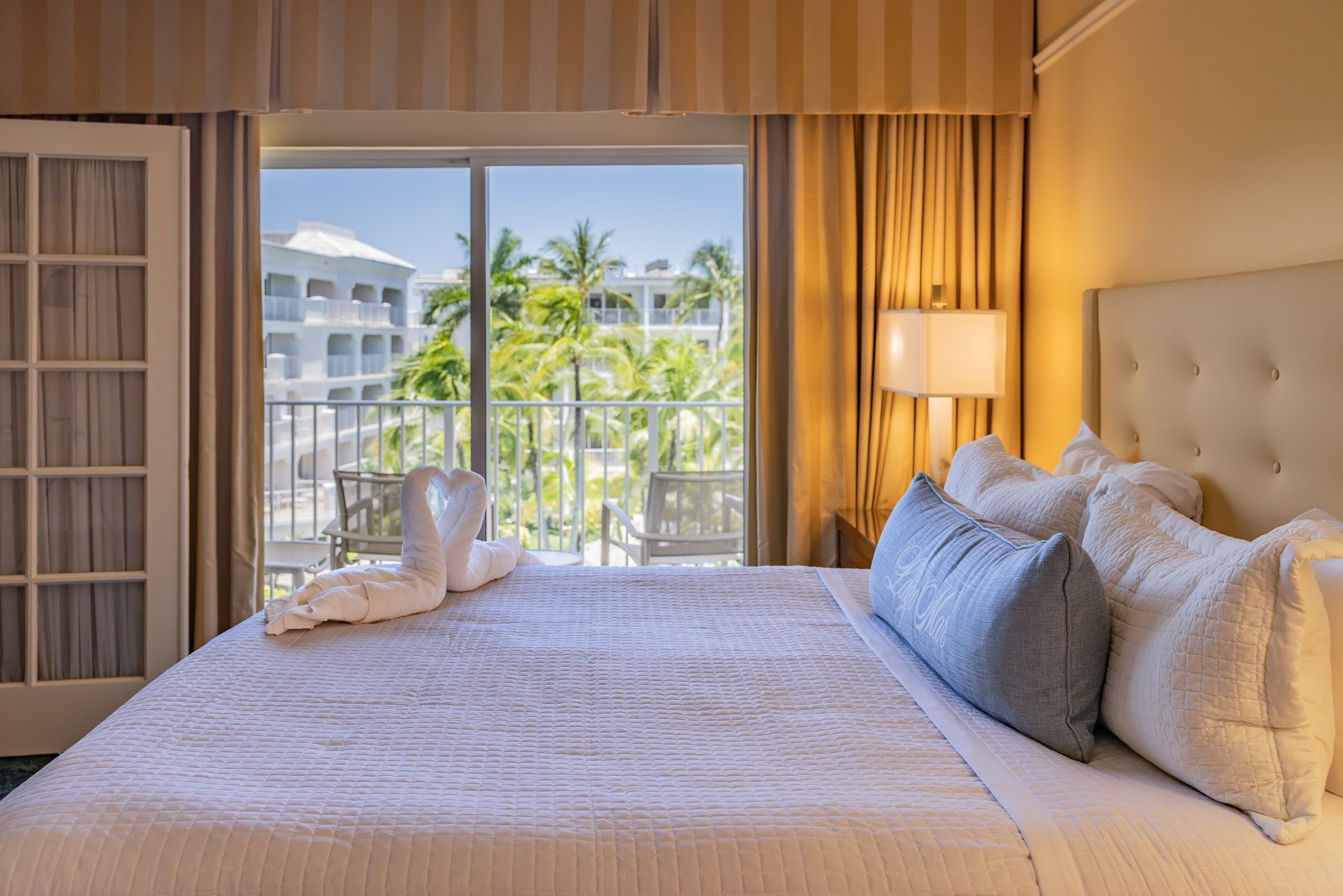 A hotel room with a large bed, a balcony with a view of palm trees and a building, and a window with curtains.