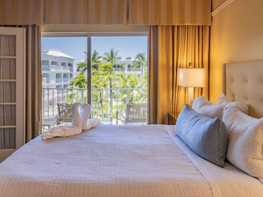 A hotel room with a large bed, a balcony with a view of palm trees and a building, and a window with curtains.
