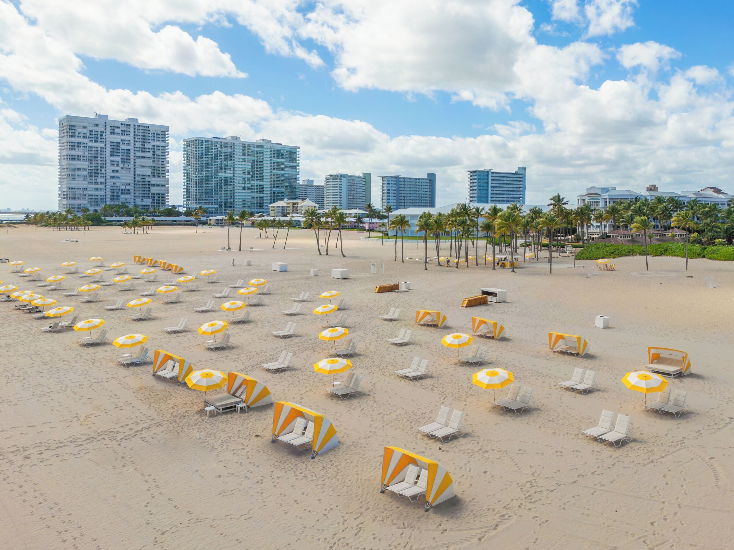 Ein Strand mit Liegestühlen und Sonnenschirmen, Palmen und Hochhäusern im Hintergrund.