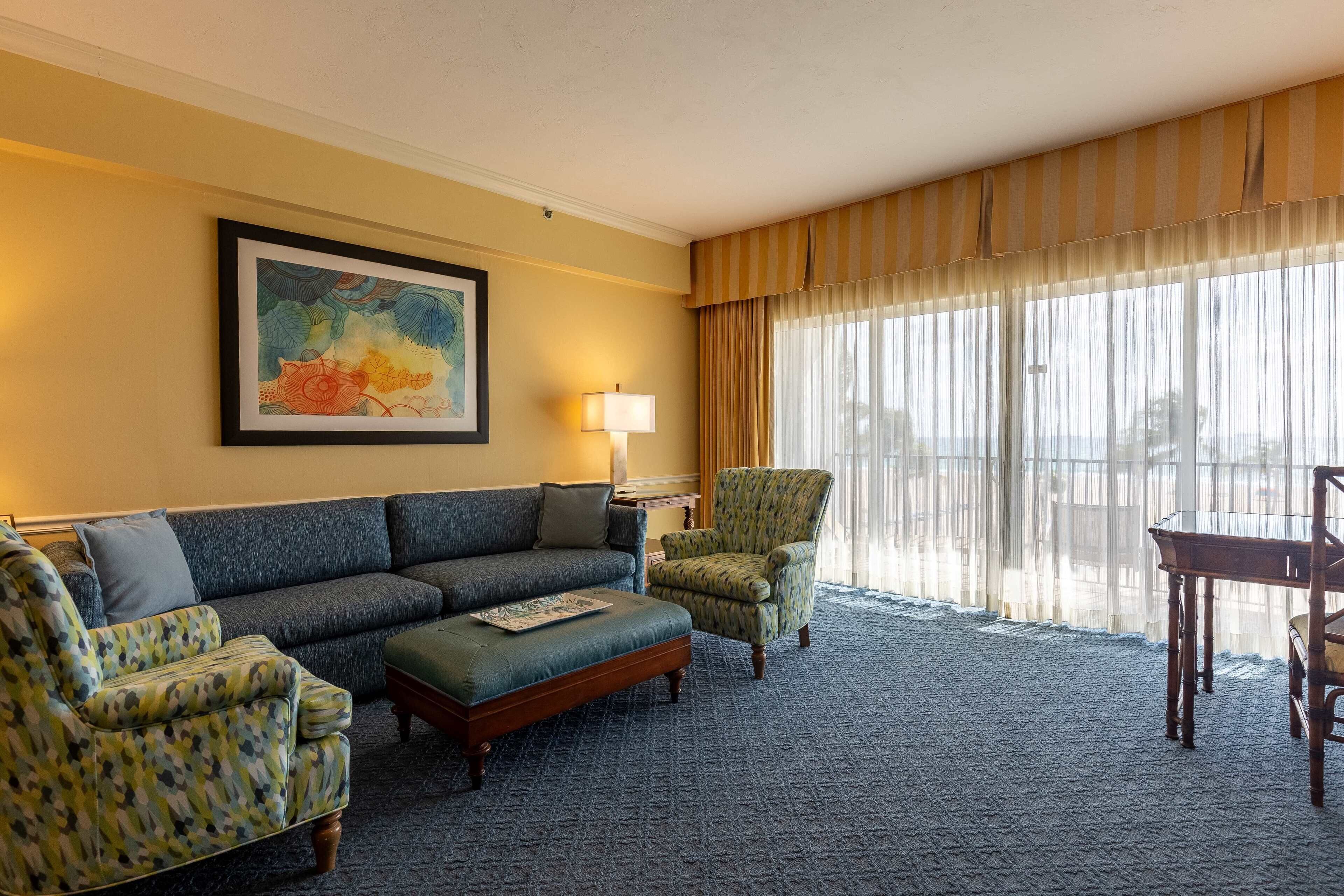 Une chambre d’hôtel avec un canapé bleu, un fauteuil à motifs, une table basse en bois et une table à manger avec des chaises. Il y a un tableau au mur et une lampe sur une table d’appoint.