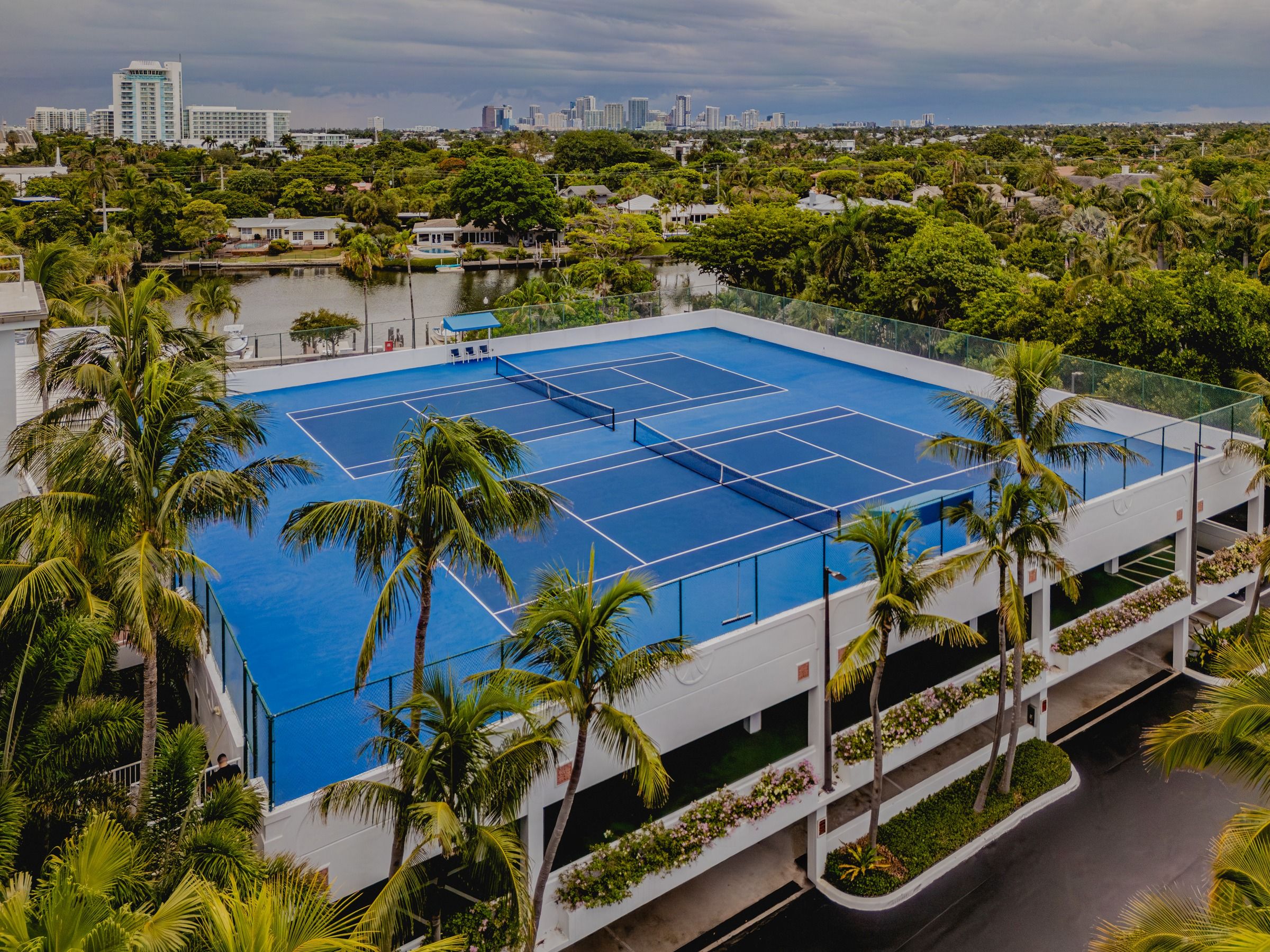 A rooftop tennis court surrounded by palm trees and a city skyline in the background.