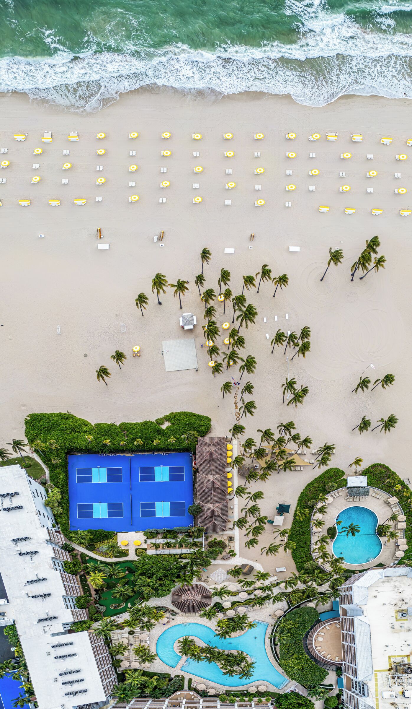 An aerial view of a beach resort with multiple sun loungers, a tennis court, a swimming pool, and palm trees.