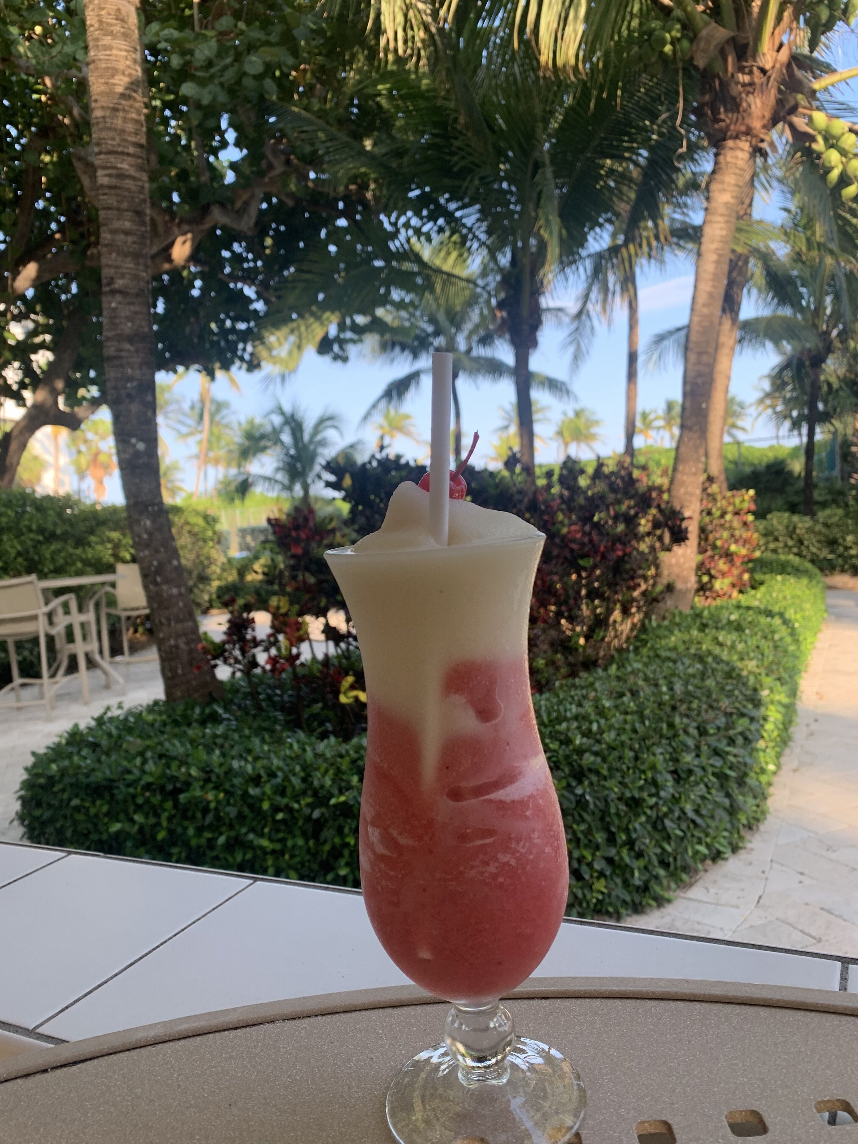 A drink in a glass with a straw, placed on a table with a view of palm trees and a patio area.
