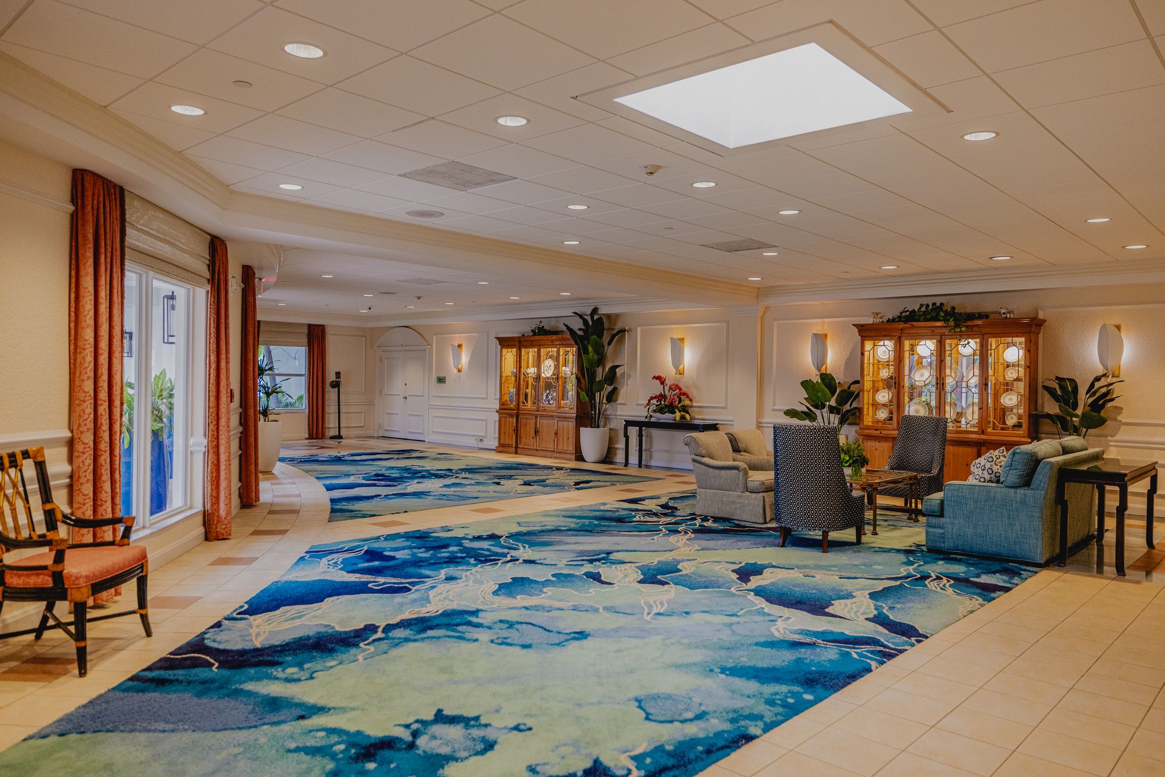 A spacious lobby with a large blue patterned carpet, a seating area with chairs and sofas, and a large wooden cabinet with glass doors.