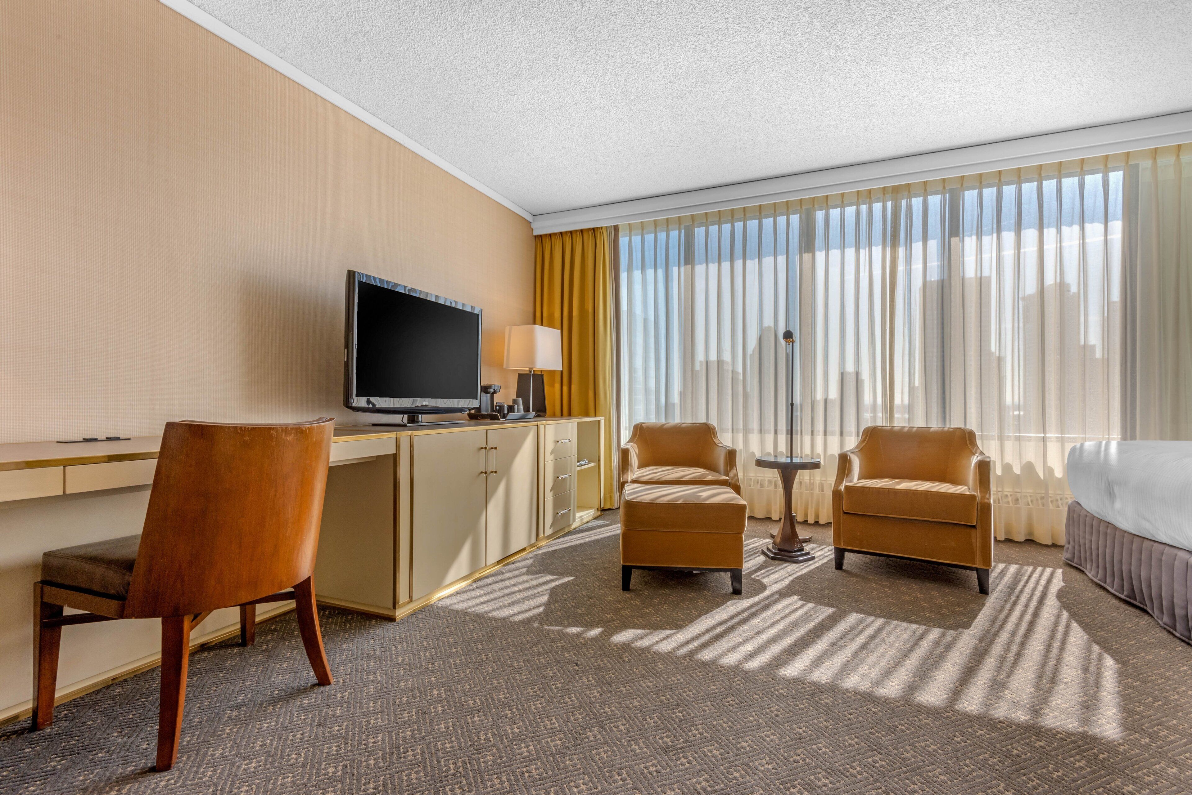 A hotel room with a desk, two armchairs, a bed, a TV, and a window with curtains.