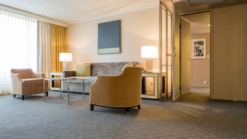 Habitación de hotel con sofá, dos sillones, una mesa de centro, un escritorio con lámpara y un cuadro en la pared.