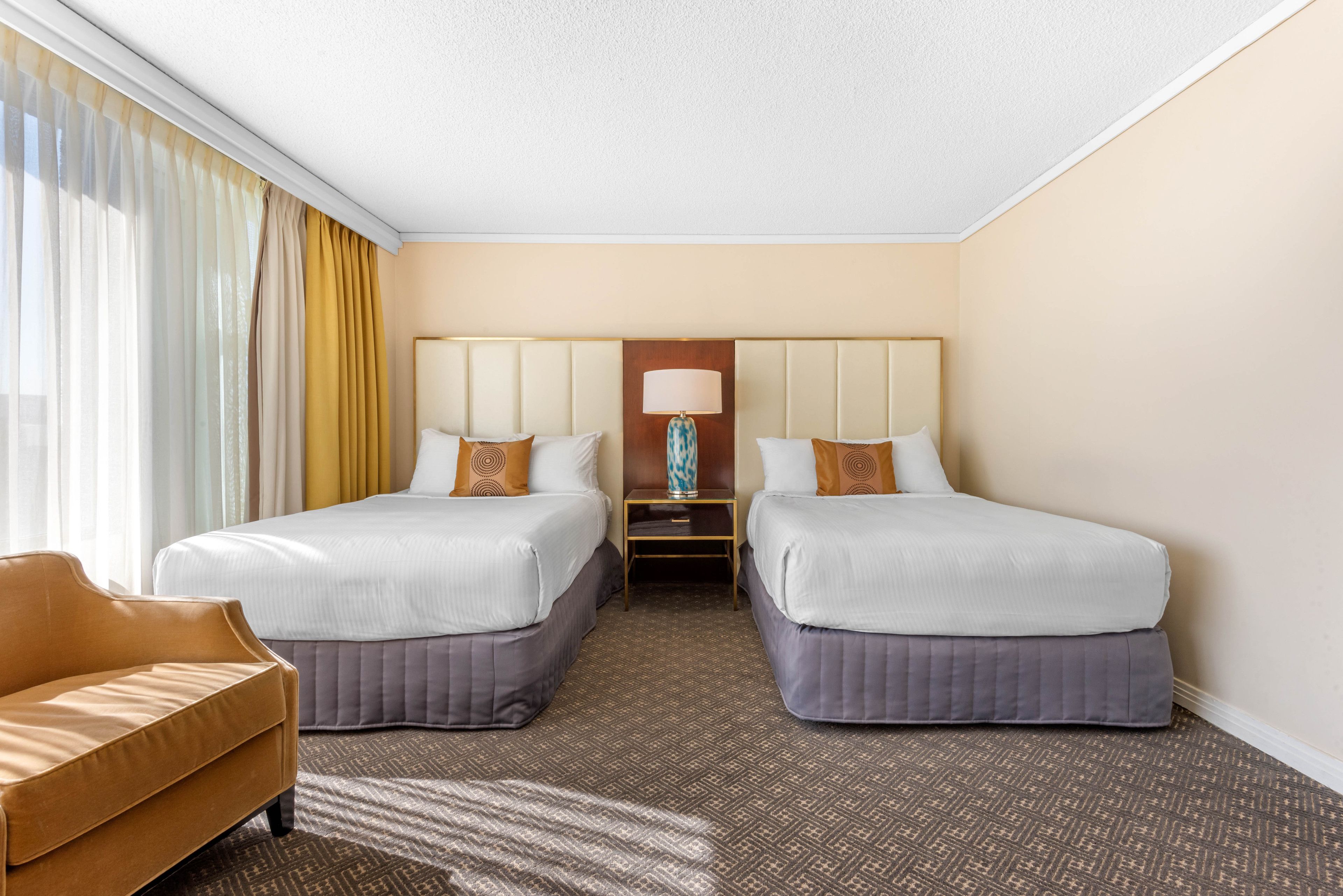 Quarto de hotel com duas camas, um sofá, uma mesinha de cabeceira com abajur e uma janela com cortinas.
