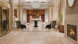 A spacious lobby with a marble floor, a central table with vases, two black armchairs, a chandelier, and a fireplace.