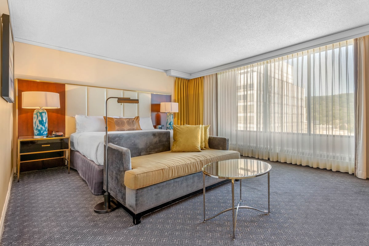 A hotel room with a sofa bed, a small table, a nightstand, and a large window with curtains.
