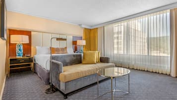 Habitación de hotel con sofá cama, una mesita, una mesita de noche y un amplio ventanal con cortinas.