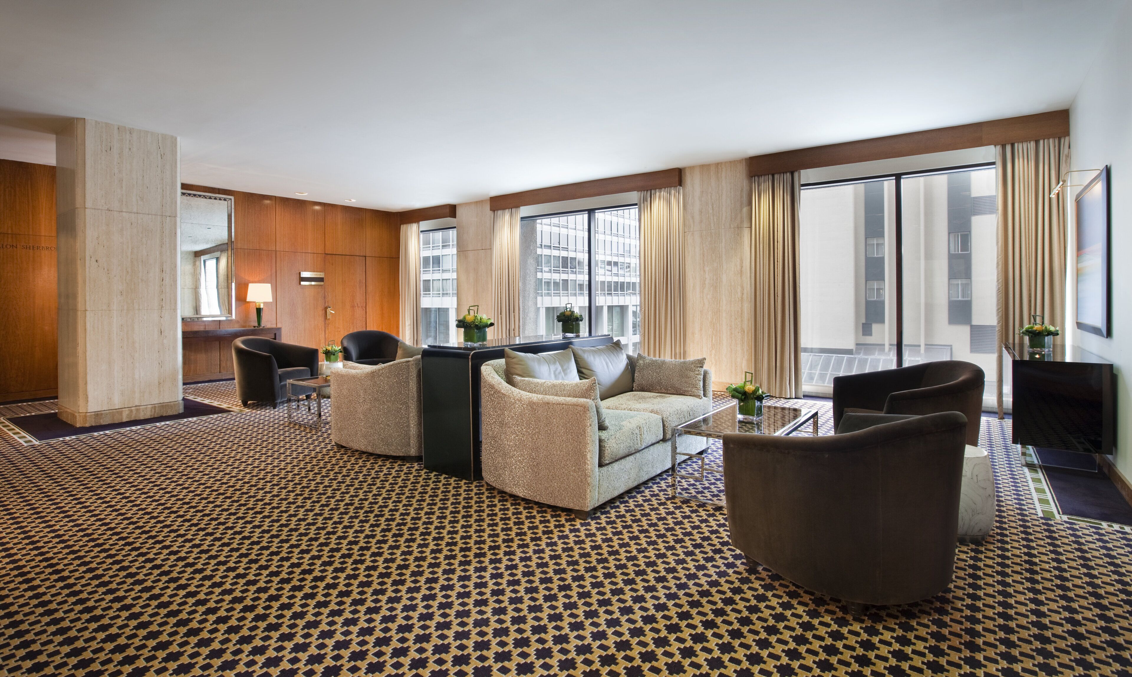 A hotel room with a patterned carpet, a sofa set, a dining table, and large windows with curtains.