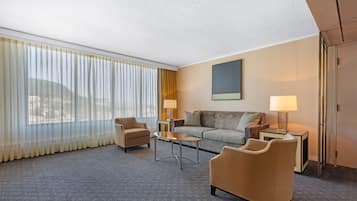 Une chambre d’hôtel avec un canapé, deux fauteuils, une table basse, une lampe et une grande fenêtre avec des rideaux.