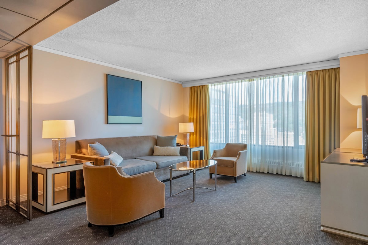 A hotel room with a sofa, armchairs, a coffee table, a TV, and a large window with curtains.