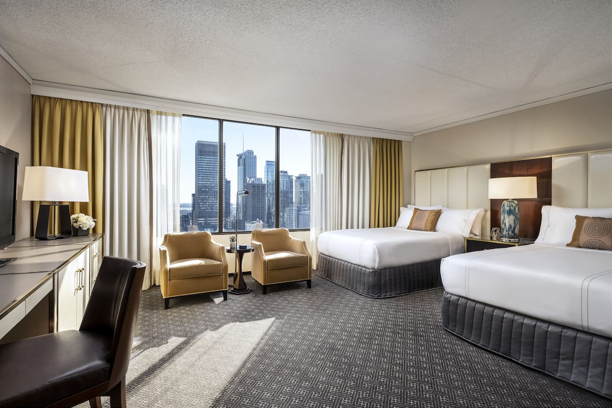 A hotel room with a large window, two beds, a desk, a chair, and a small table.
