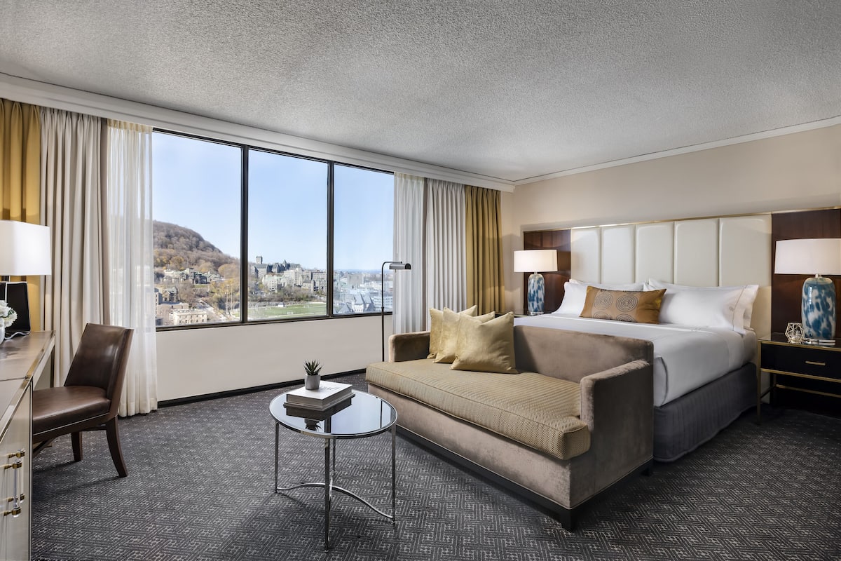 A hotel room with a large bed, a sofa, a desk, and a view of a cityscape through large windows.