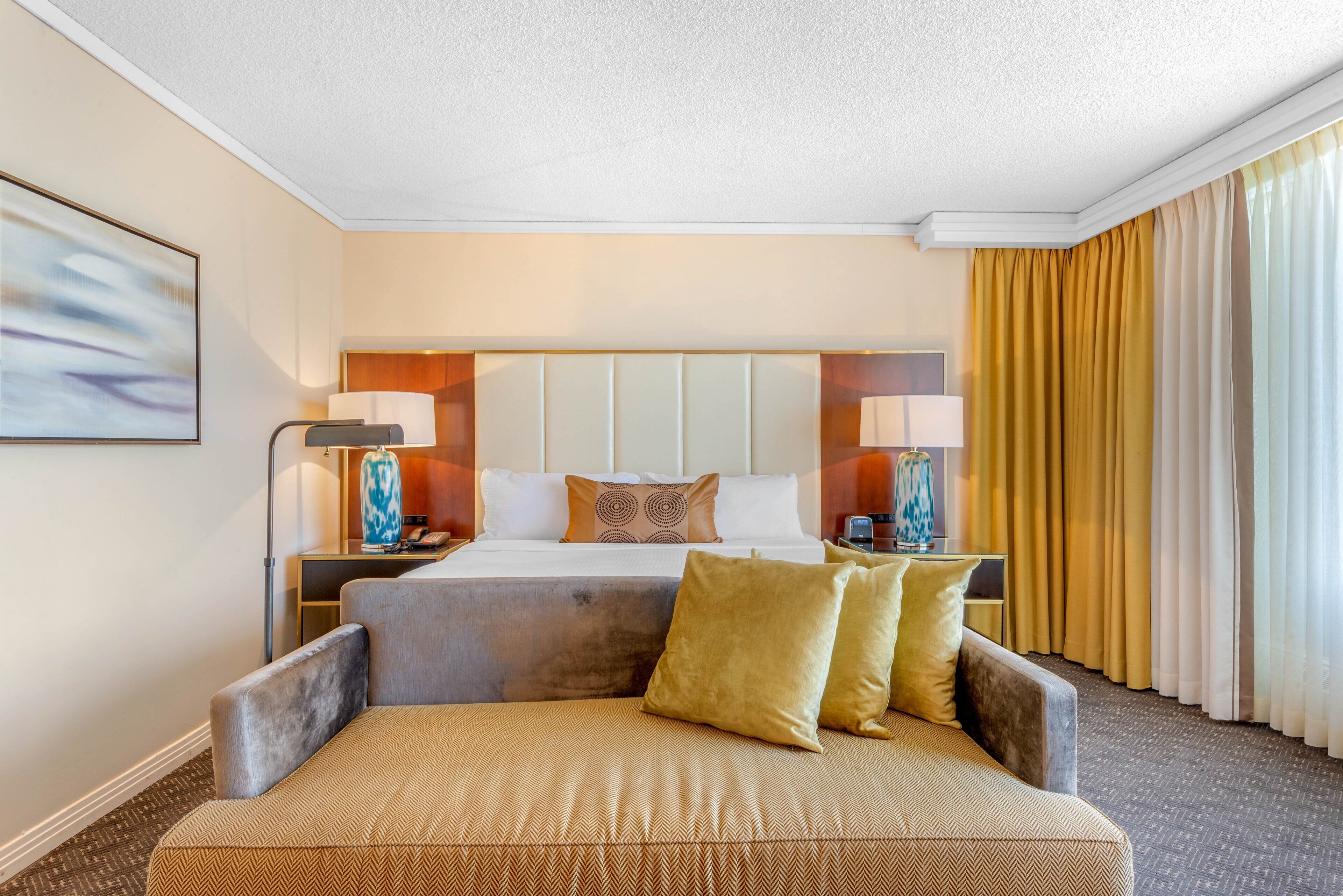 A hotel room with a large bed, two bedside tables with lamps, a sofa, and a large window with curtains.