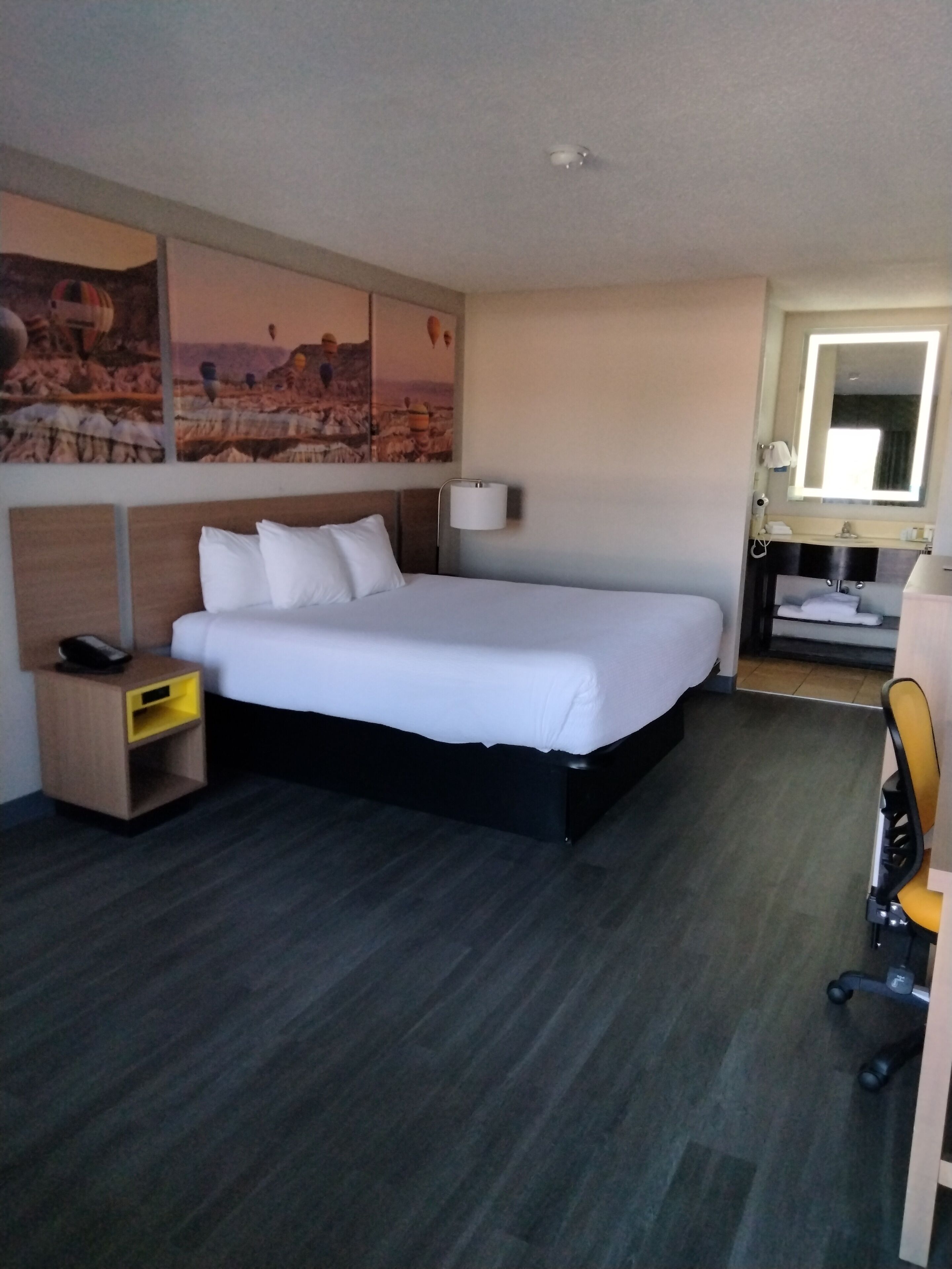A hotel room with a large bed, a bedside table with a phone, a small desk with a chair, a mirror, and artwork on the wall.
