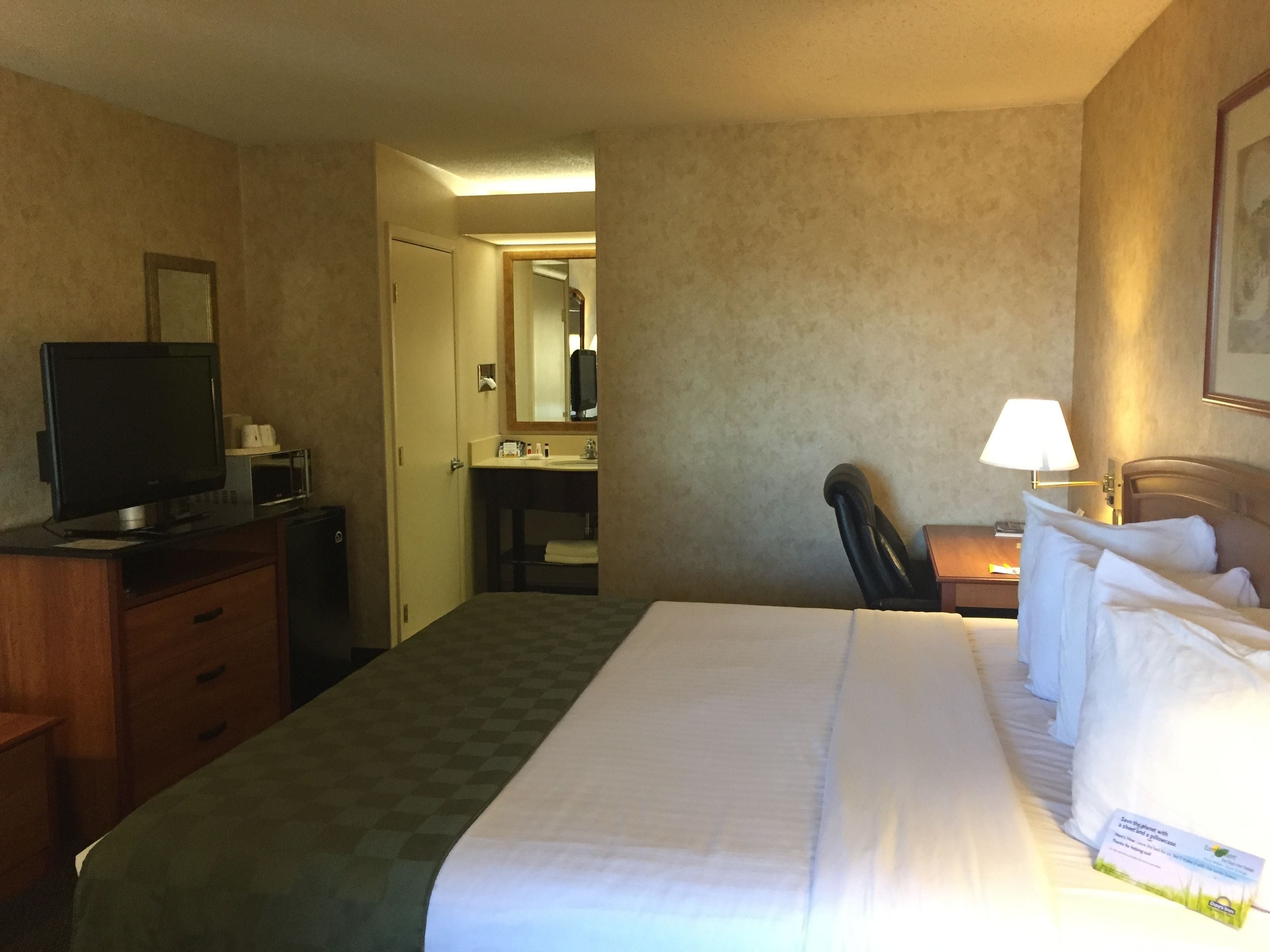 A hotel room with a bed, a desk with a computer, a television, a chair, and a bathroom visible through an open door.