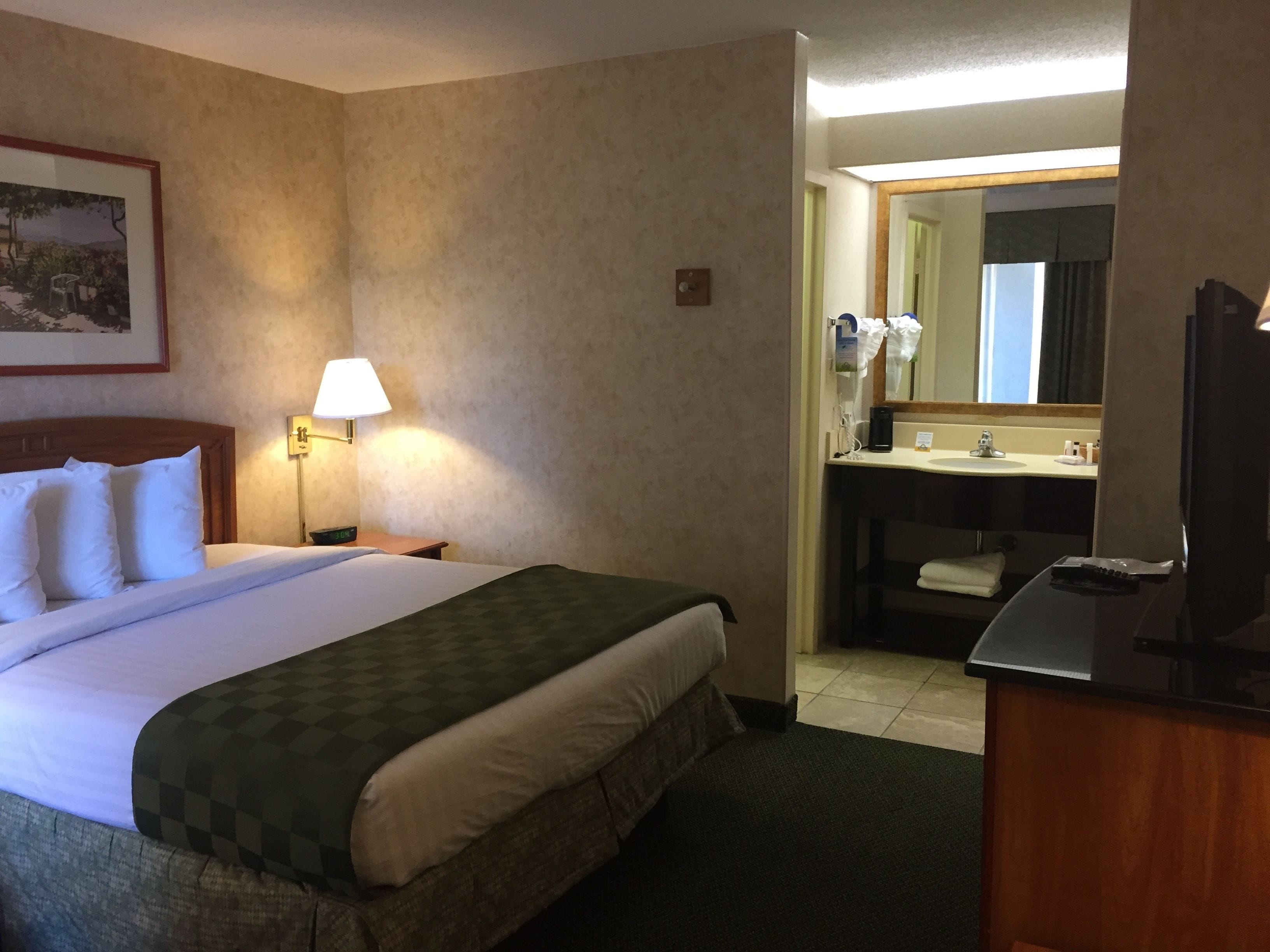 Habitación de hotel con cama, mesita de noche con lámpara, baño con lavamanos y espejo, y escritorio con silla.