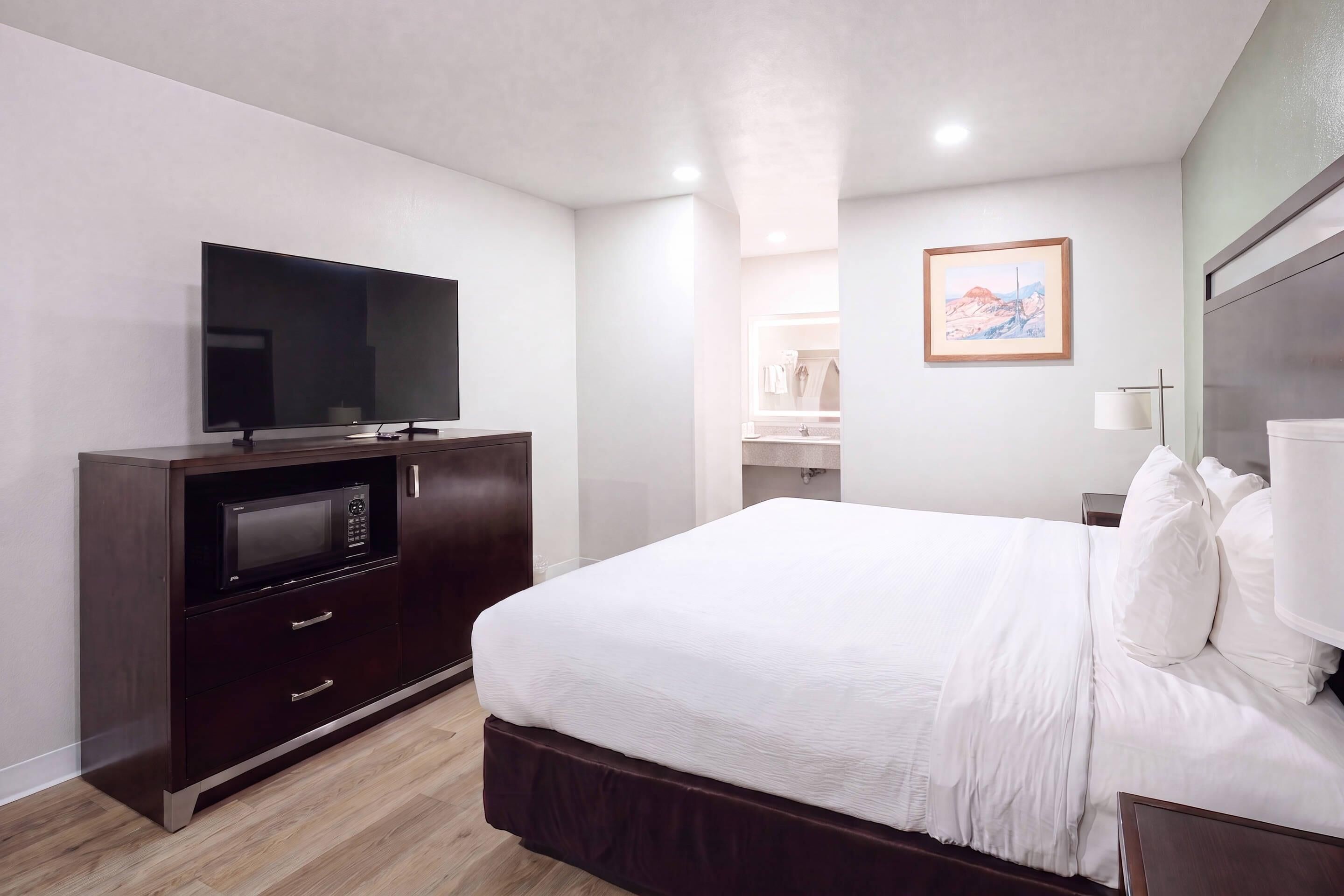 A hotel room with a bed, a television, a microwave, a painting, and a bathroom visible through an open door.