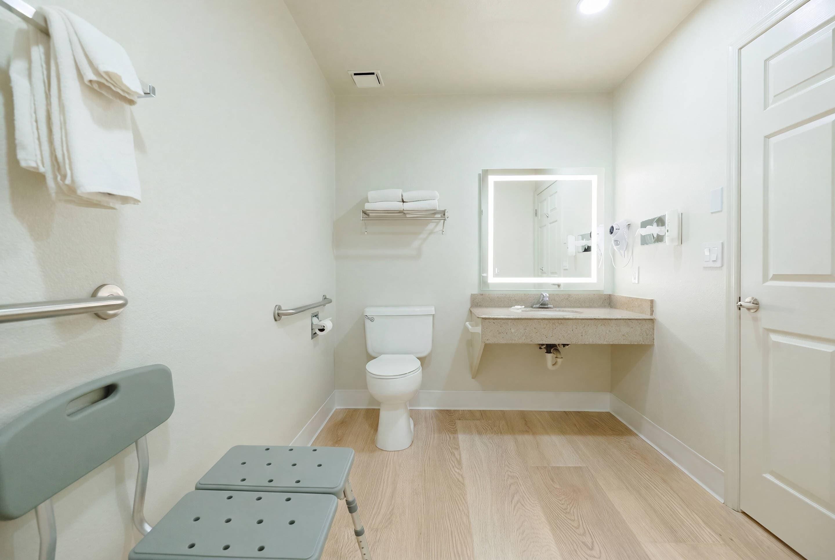 A small, clean bathroom with a toilet, sink, and a mirror above the sink. There is a towel rack on the wall and a step stool in the corner.