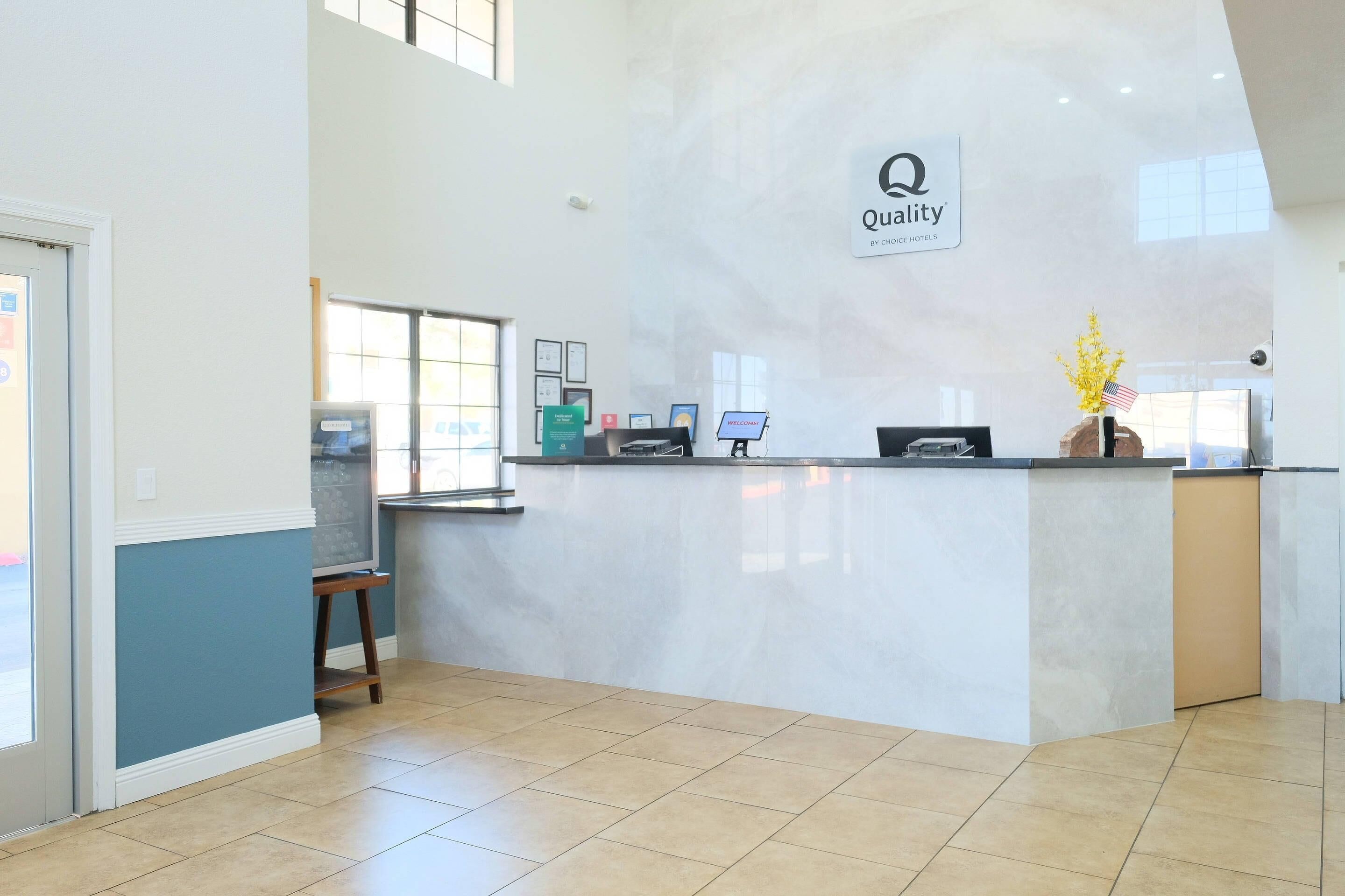 Uma área de recepção de hotel com balcão de mármore, um aviso indicando 'Quality by Choice Hotels', um computador e uma pequena mesa com uma cadeira.
