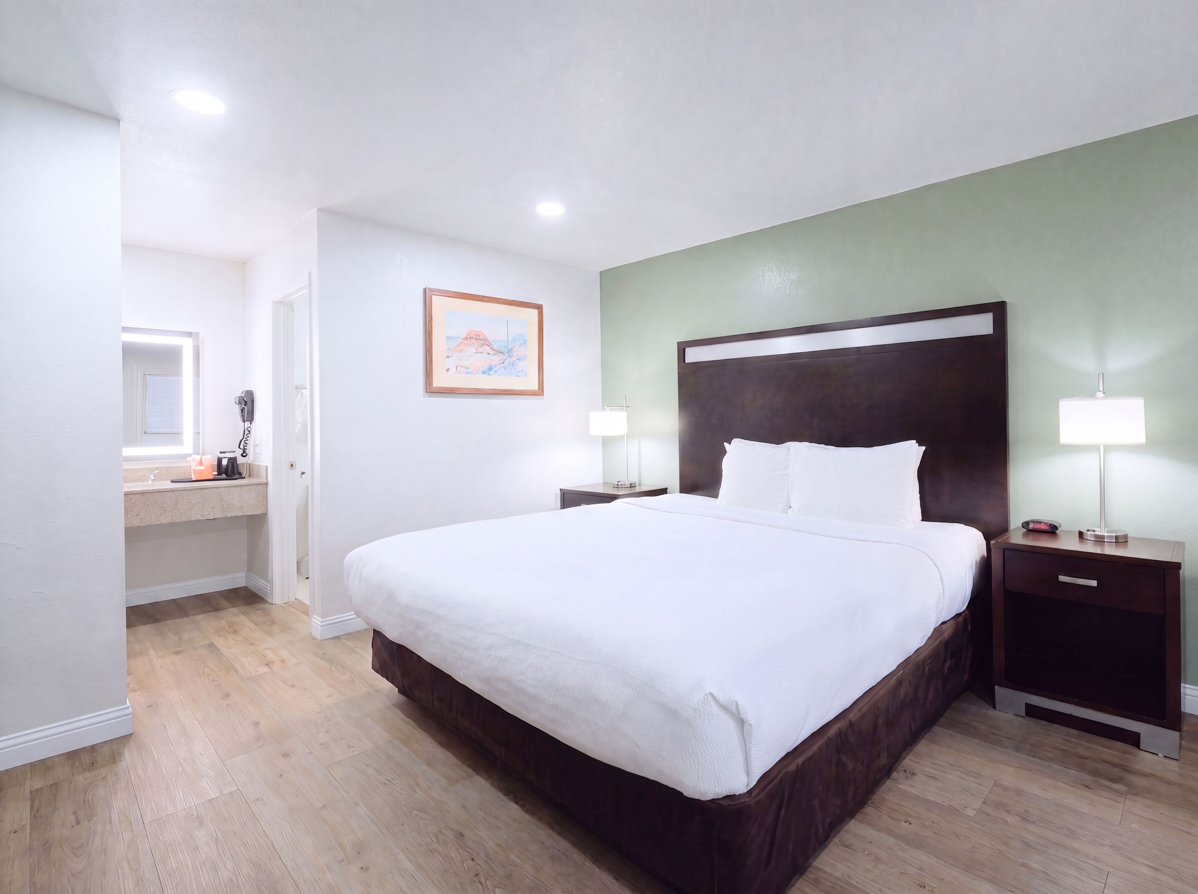 Quarto de hotel com uma cama grande, mesas de cabeceira, um espelho, um quadro na parede e piso de madeira.