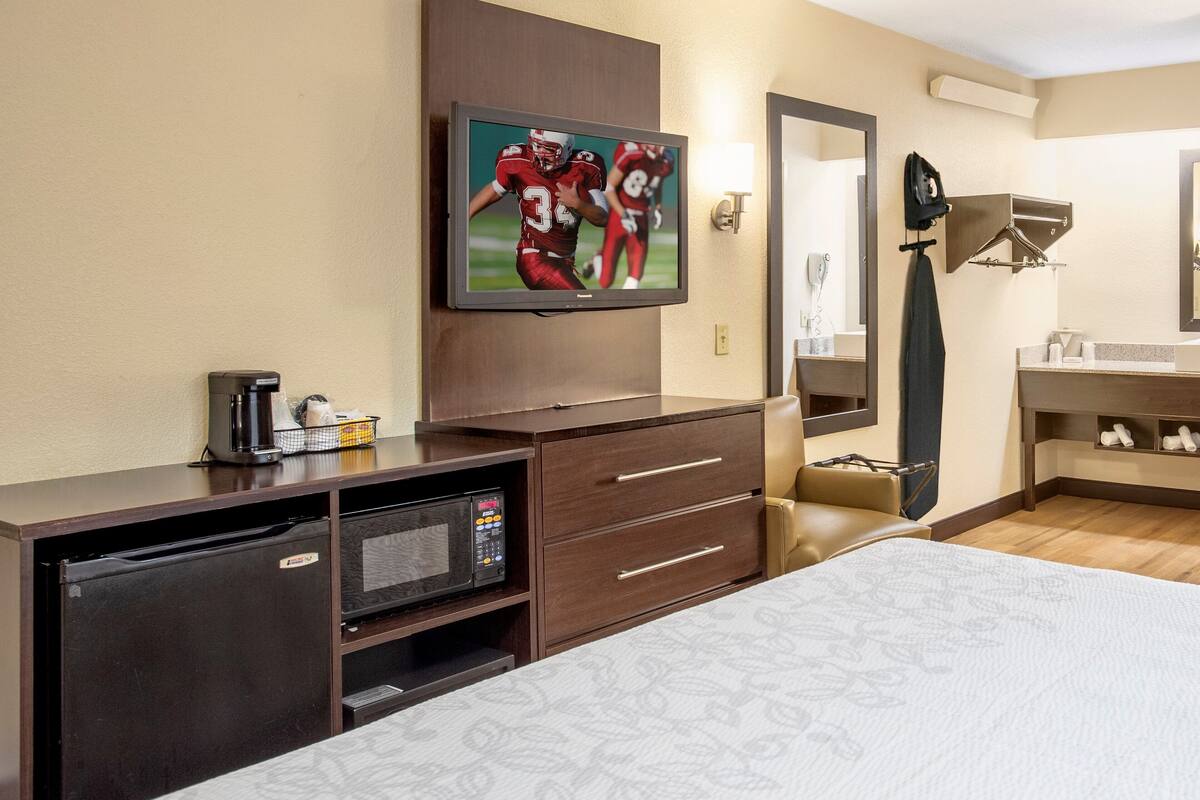 A hotel room with a bed, a TV mounted on the wall, a desk with a coffee maker, a microwave, and a mirror.