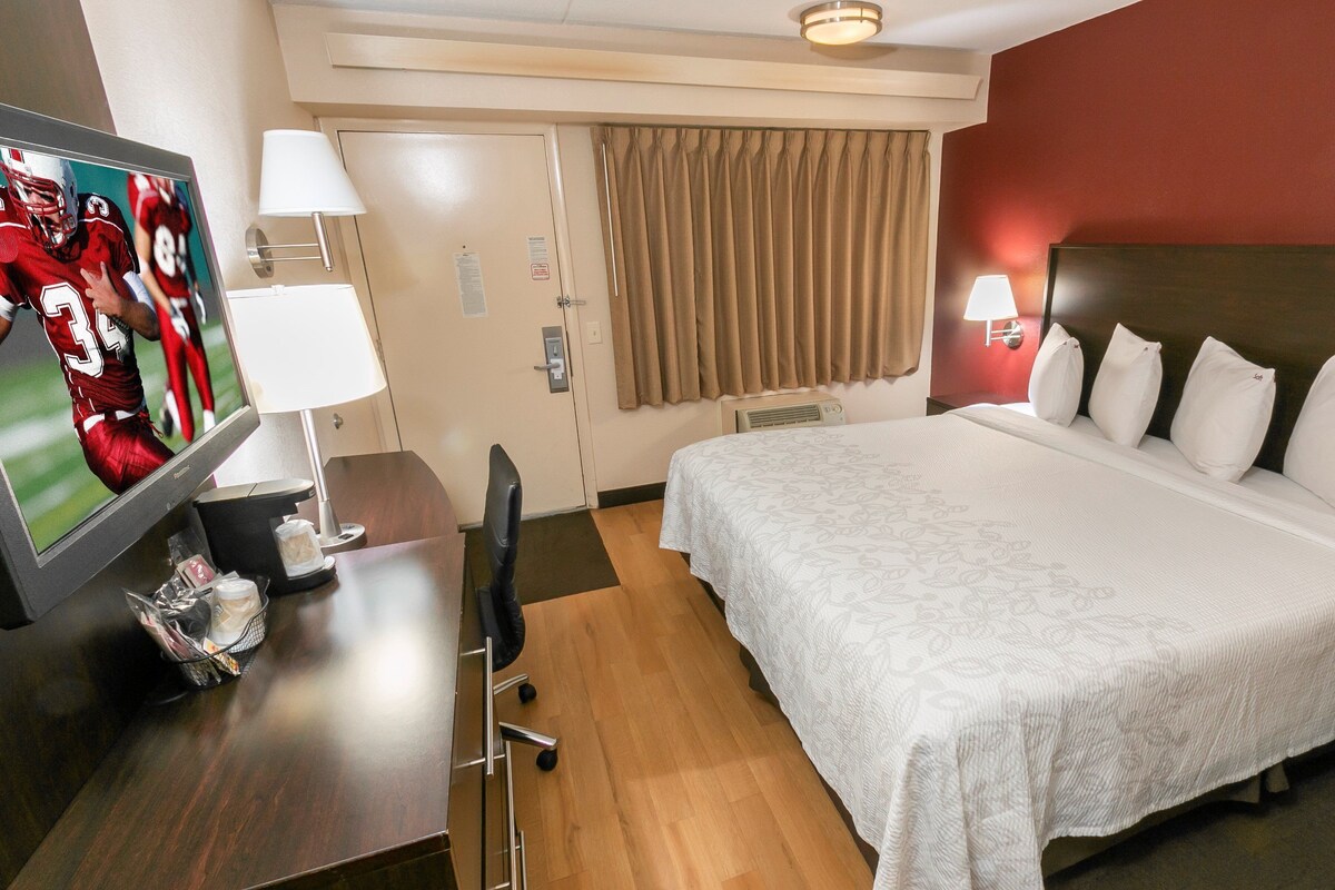 Habitación de hotel con cama, escritorio con televisor, una silla, lámpara y ventana con cortinas.