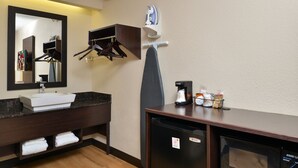 Baño de hotel con lavamanos, espejo, tabla de planchar y un refrigerador compacto.