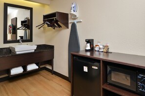 Baño de hotel con lavamanos, espejo, tabla de planchar y un refrigerador compacto.