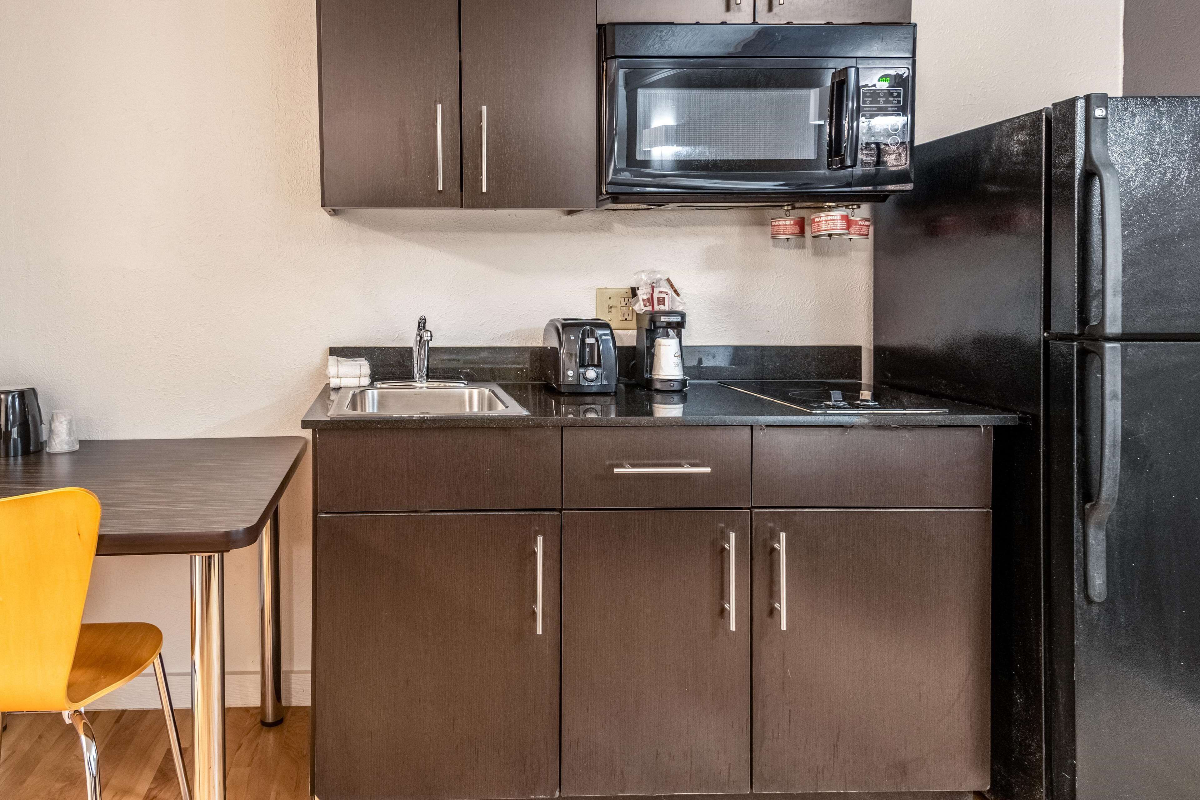 A compact kitchenette with dark brown cabinets, a black microwave, a black refrigerator, a sink, a toaster, and a coffee maker.